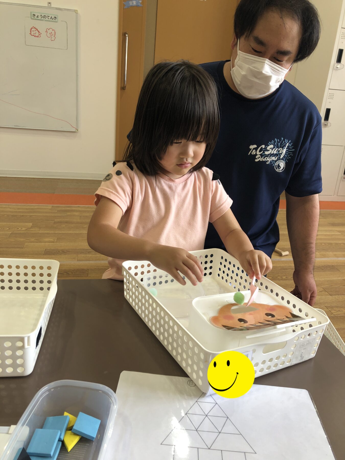 Young girl in a pink shirt works on a craft with blue foam blocks as a masked man stands beside her in a classroom setting.