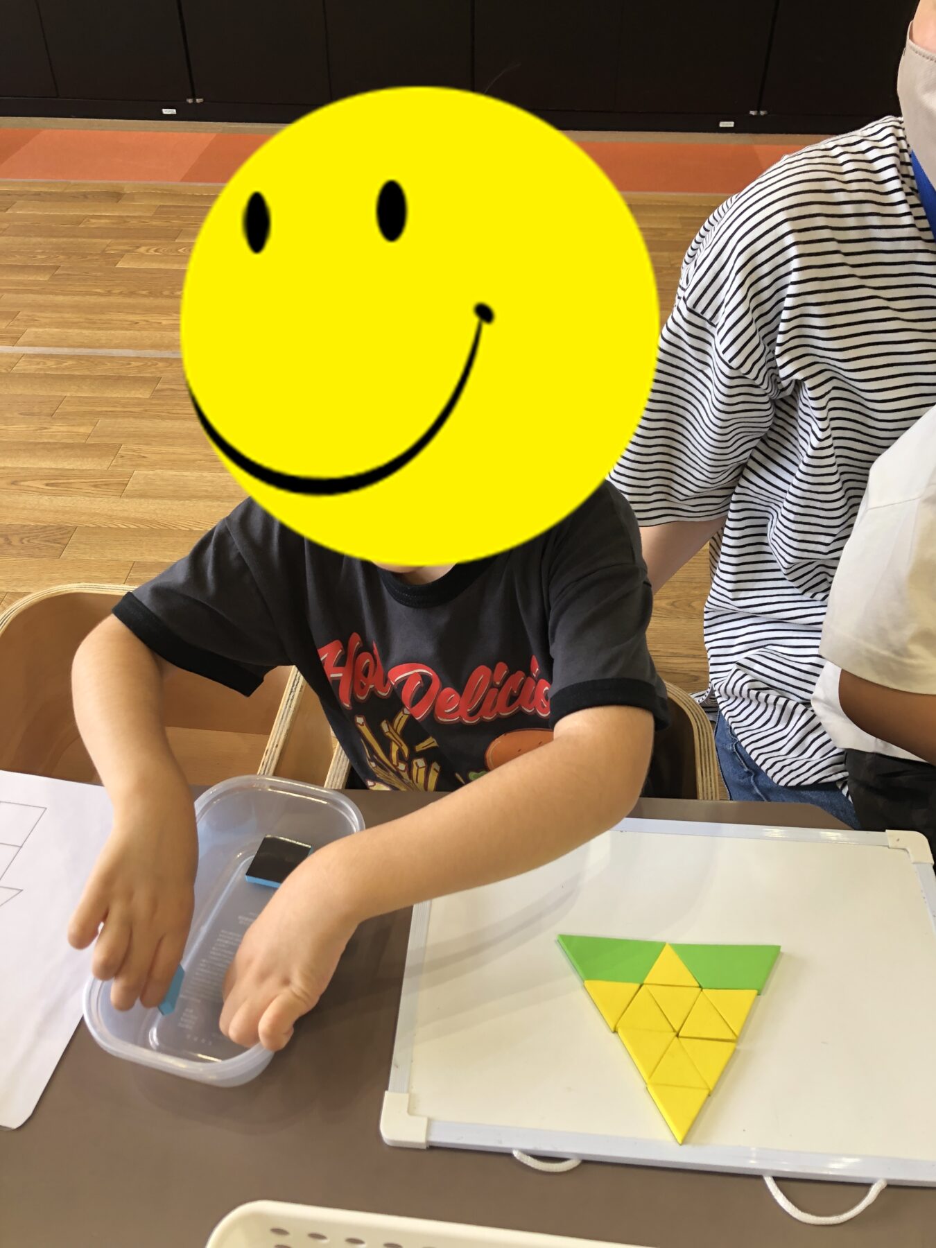 Child sits at a table assembling a yellow-and-green triangular puzzle on a white tray; a large yellow smiley sticker covers their face for privacy.