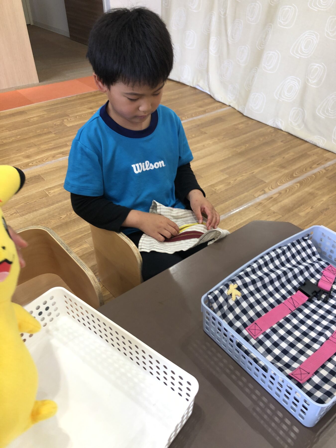 Young boy in a bright blue Wilson shirt sits at a table, folding striped fabric for a craft project in a classroom or playroom.