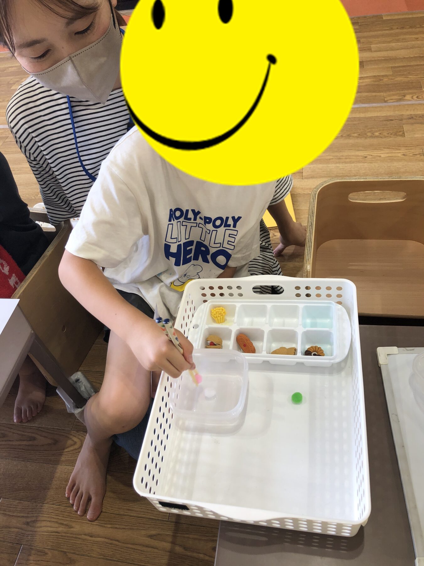Child at a hands-on activity tray with small colored pieces, supervised by an adult wearing a mask nearby.