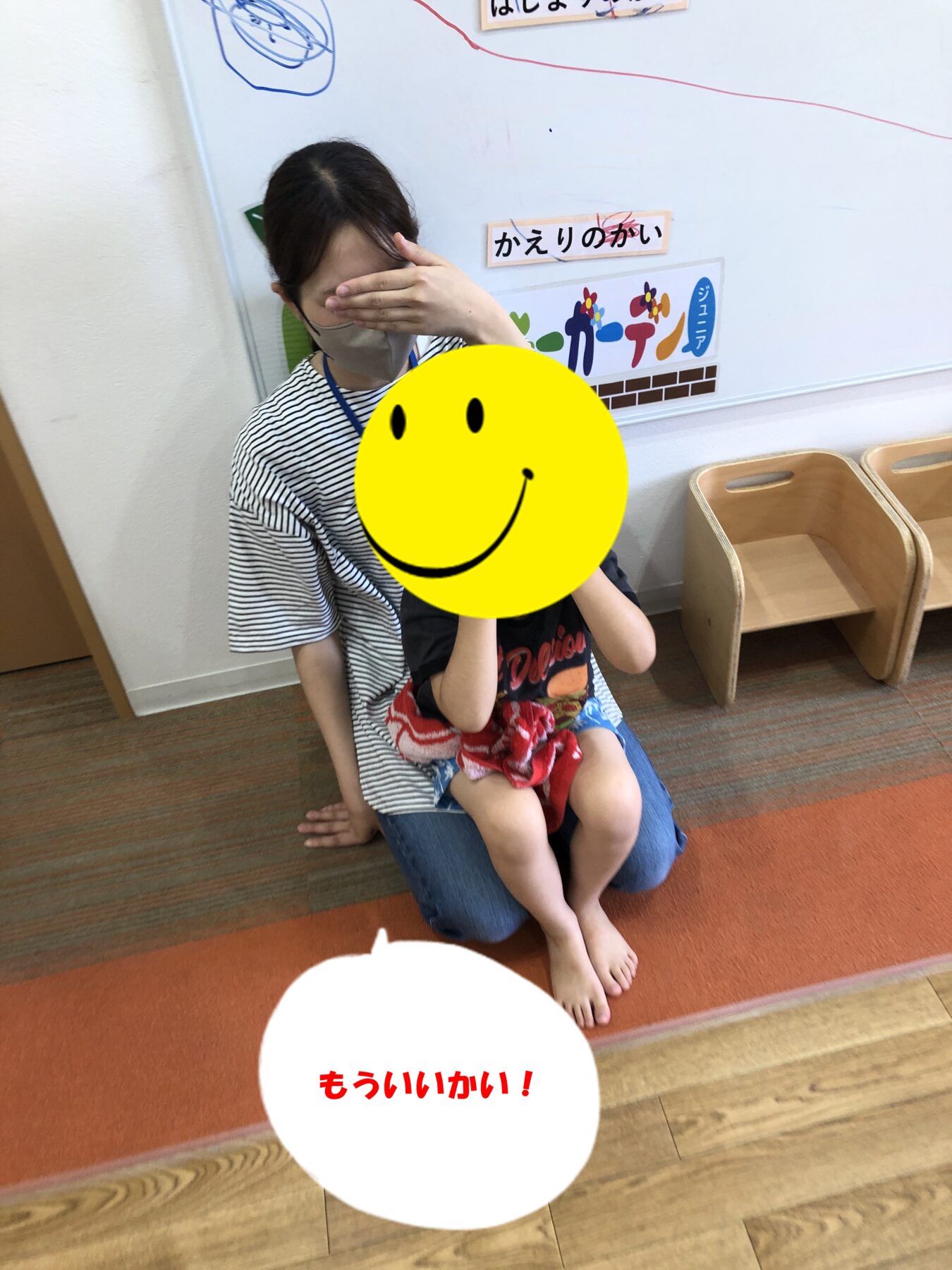 Adult woman in a striped shirt sits on the floor with a child on her lap; the woman covers her eyes with her hand, while a large yellow smiley sticker obscures part of their faces in a classroom setting.