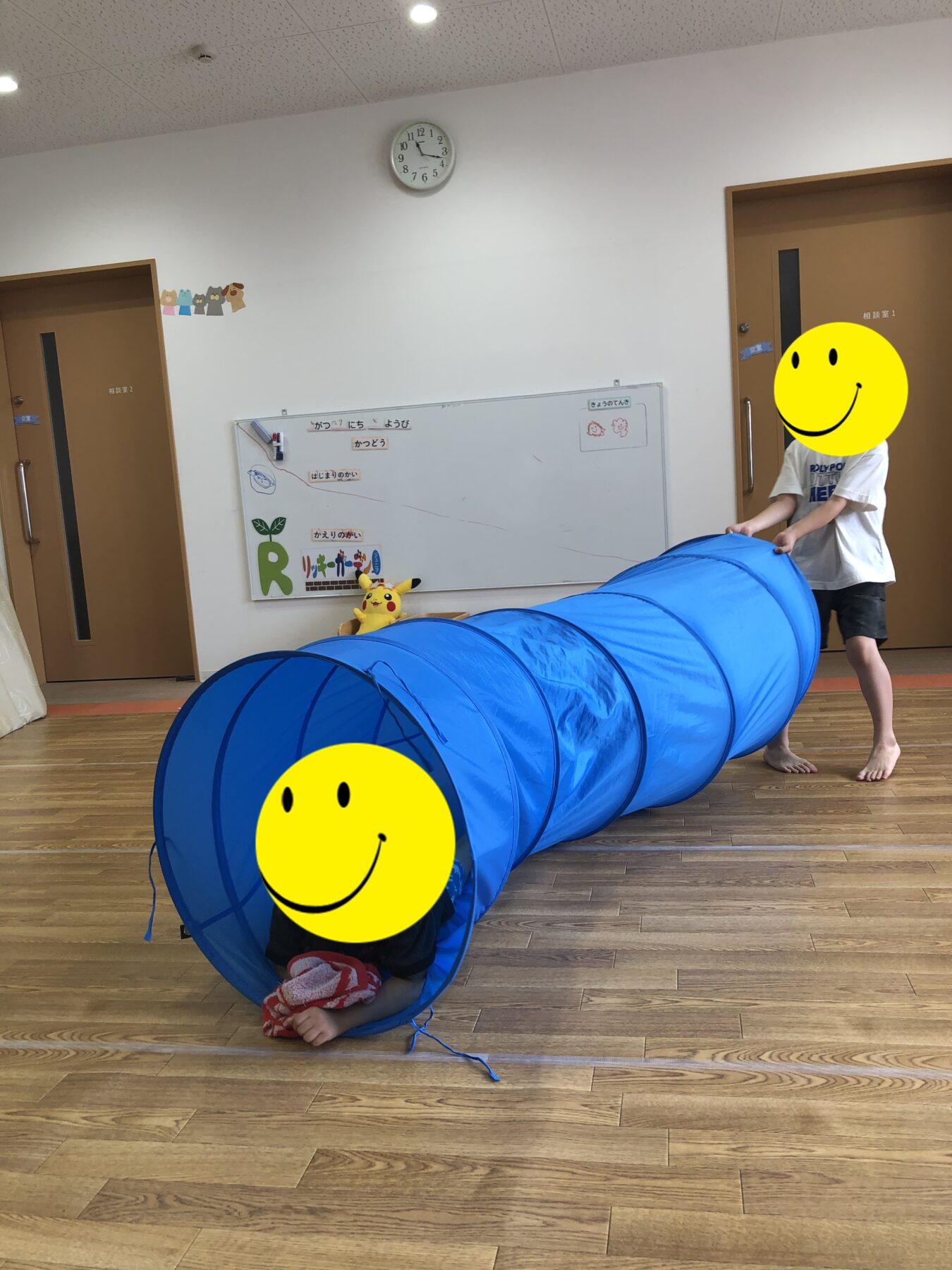 Child pushes a bright blue fabric play tunnel along a wooden floor in a classroom or playroom; another child sits inside the tunnel at the front.