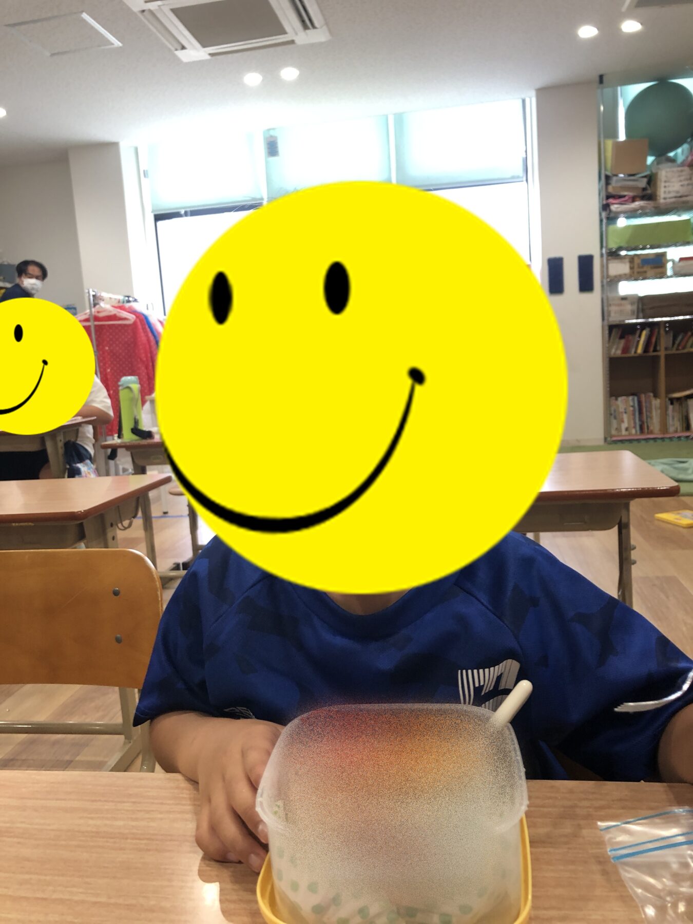 Child in a blue shirt sits at a desk in a classroom, a large yellow smiley sticker covers their face for privacy.