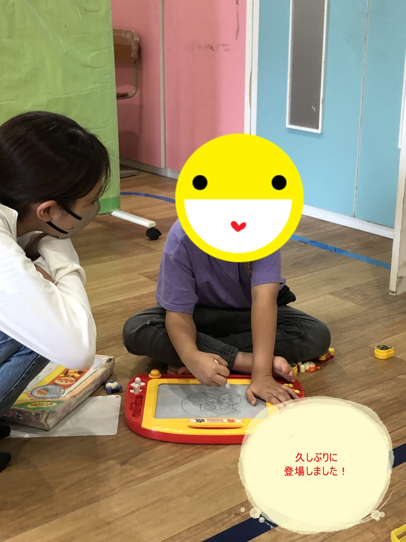 Two children sit on a wooden floor, playing with a red-and-yellow electronic drawing tablet; colorful room around them.