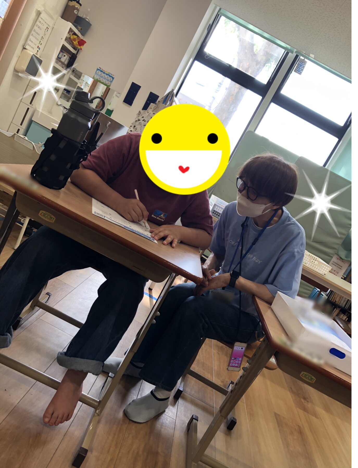 Student writes at a desk while a staff member in a mask sits nearby in a classroom with a large window behind.