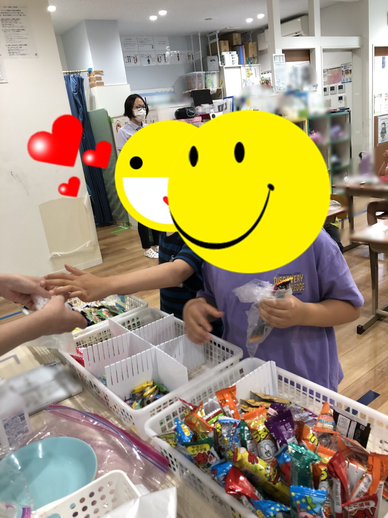 Children participate in a candy distribution activity in a bright room; baskets with assorted snacks on a table, a child hands a treat to someone off-frame while a staff member stands in the background.