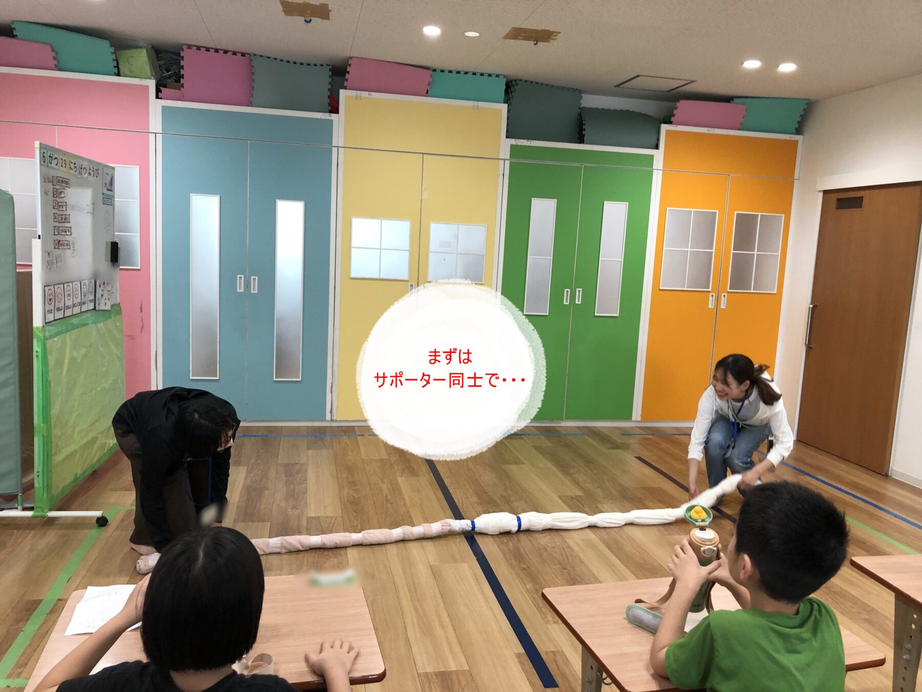 Bright classroom/gym with rainbow lockers (blue, yellow, green, orange), mats on the wall, and a long rope on the floor for a group activity; students sit at desks nearby; instructor kneels off to the side.