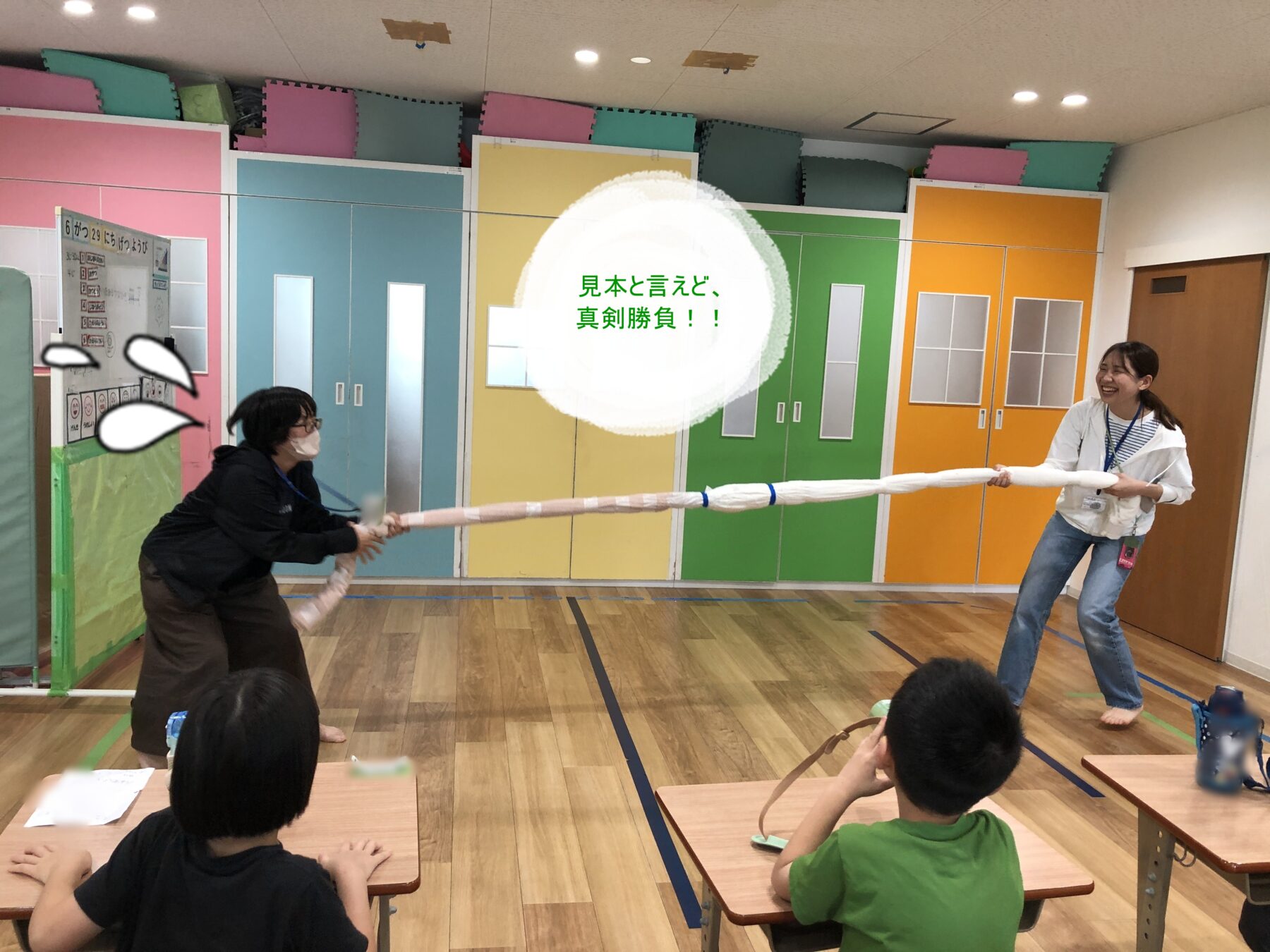 Two adults compete in a tug-of-war with a long rope in a colorful classroom while children sit at desks and watch. A white circular overlay with green Japanese text sits in the center.