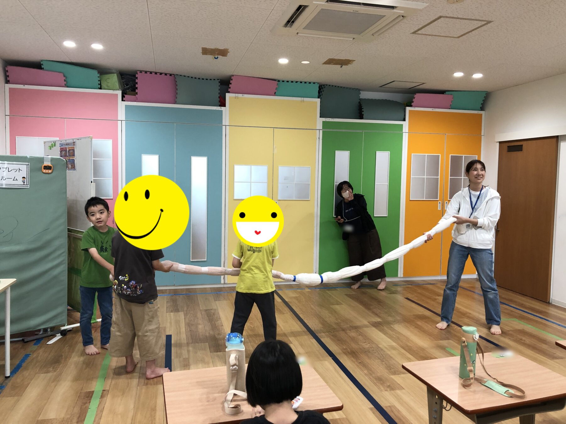 Children and adults hold a long white fabric rope in a colorful classroom, playing a game together in a gym-like room.