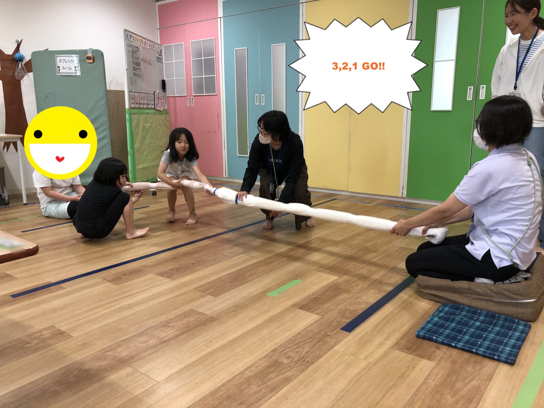 Children and adults tug a long white cloth rope in a colorful gym, pulling together in a group activity.