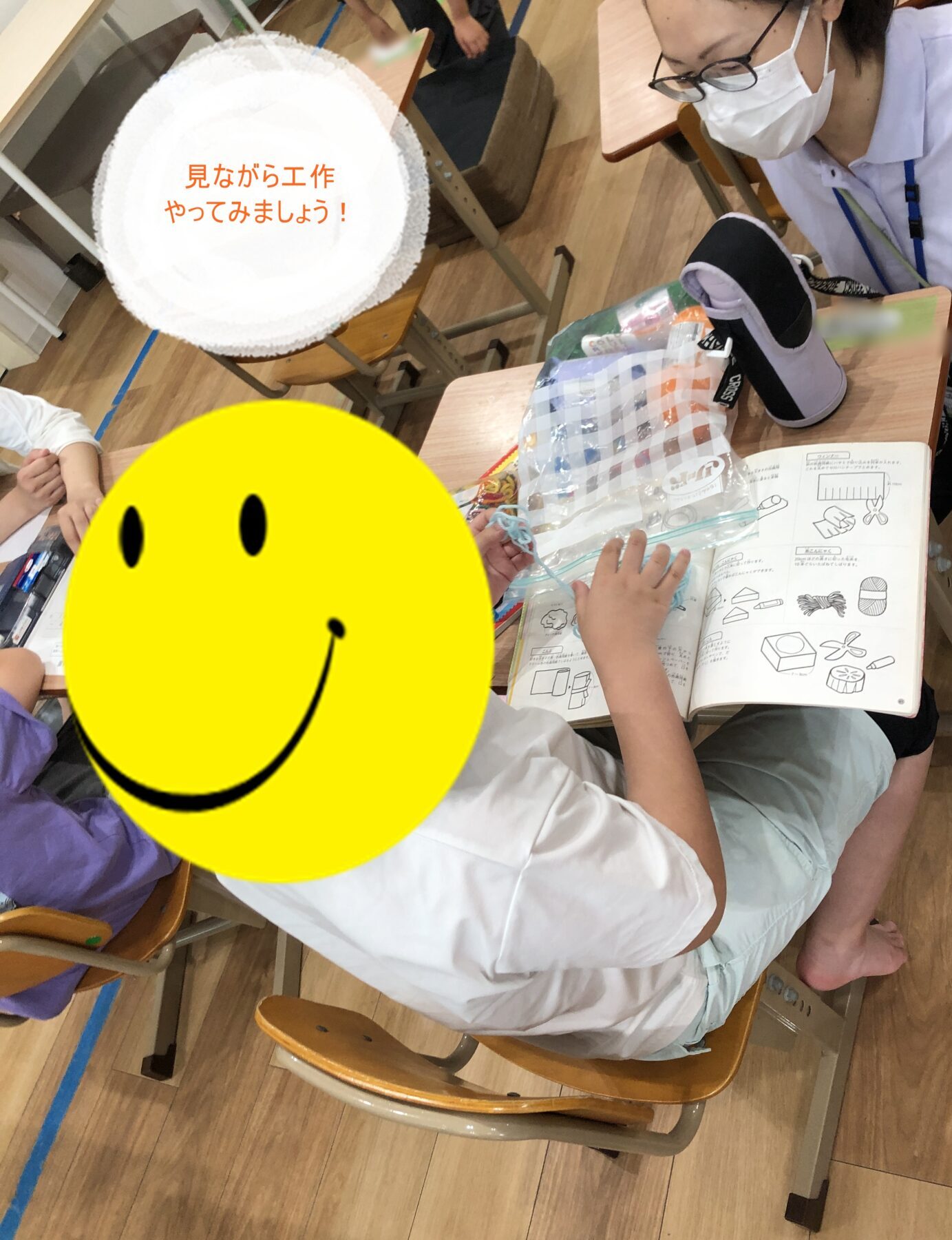 Classroom scene of students doing a craft activity at desks with an instruction book and supplies nearby; a teacher wearing a mask oversees the activity.