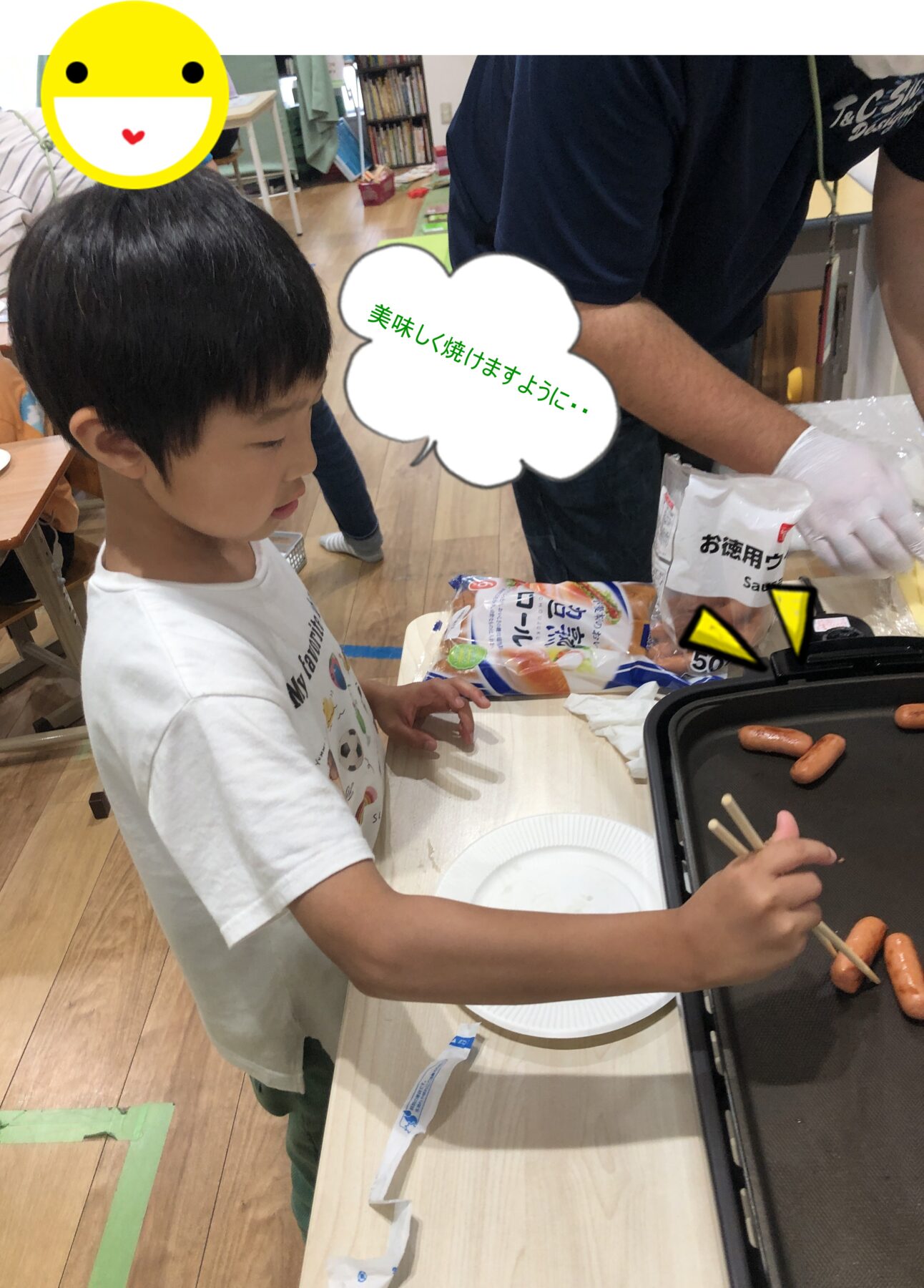 Child grilling sausages on a hot plate with tongs, assisted by an adult indoors; speech bubble in Japanese says '希望...' or similar (translation: '...')