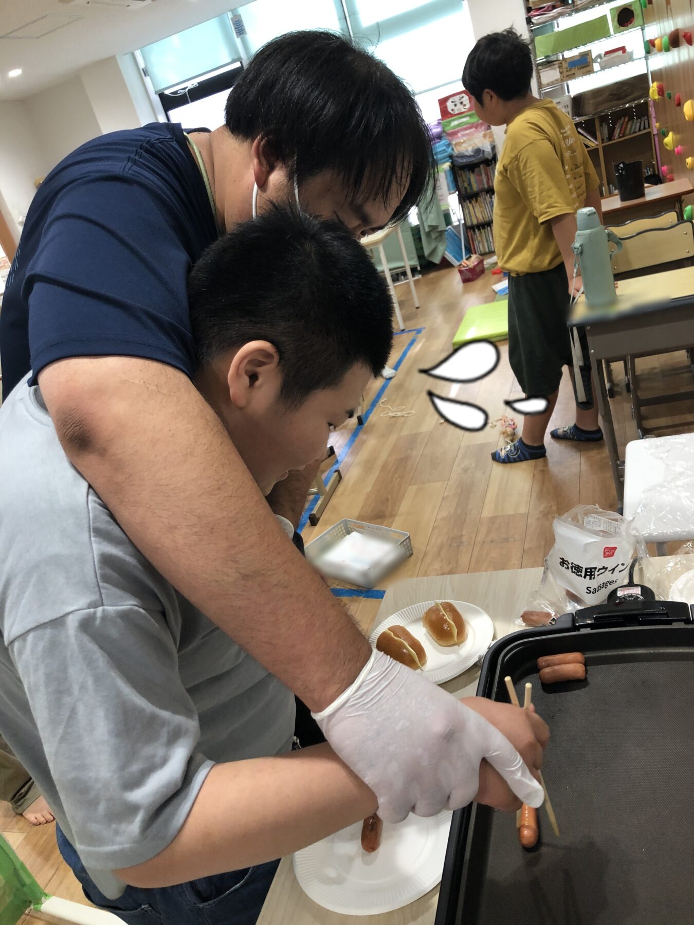 A man wearing gloves ties hot dogs on a grill while a child sits nearby with buns on a plate in a classroom kitchen setting.