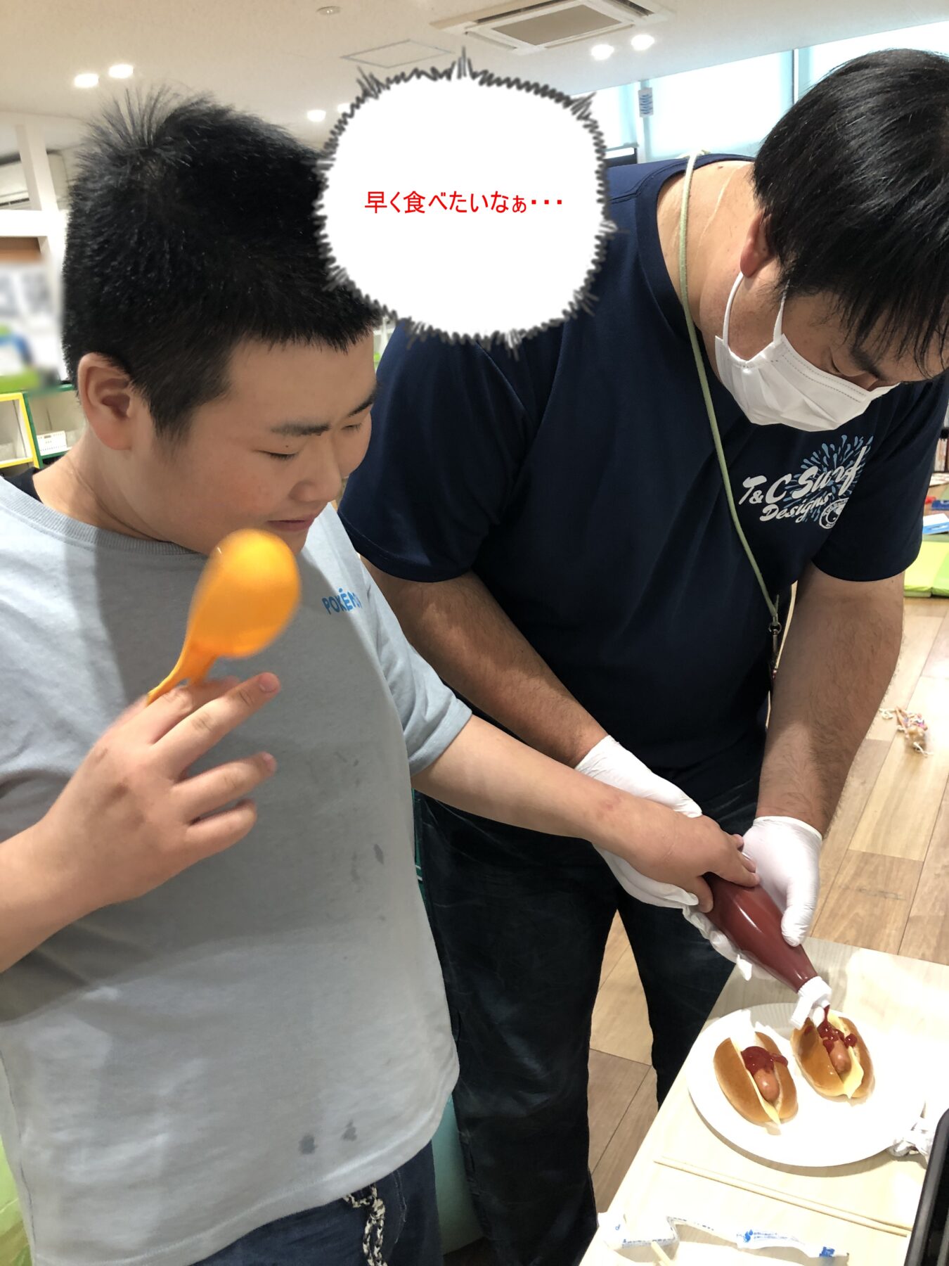Two people in a bright kitchen: a boy holding an orange spatula while a masked adult assists with preparing hot dogs and toppings on a table.