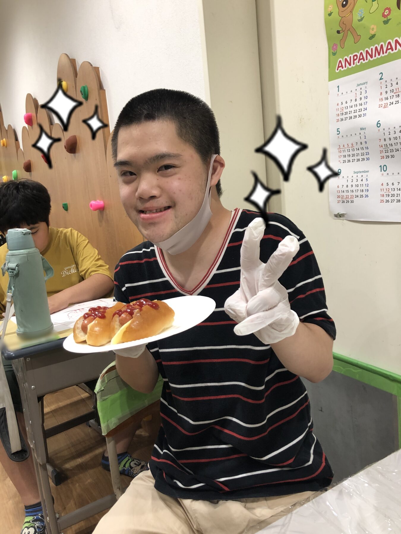 Smiling young man in a black and red striped shirt holds a plate with a hot dog topped with ketchup, wearing a mask under his chin in a casual dining area.