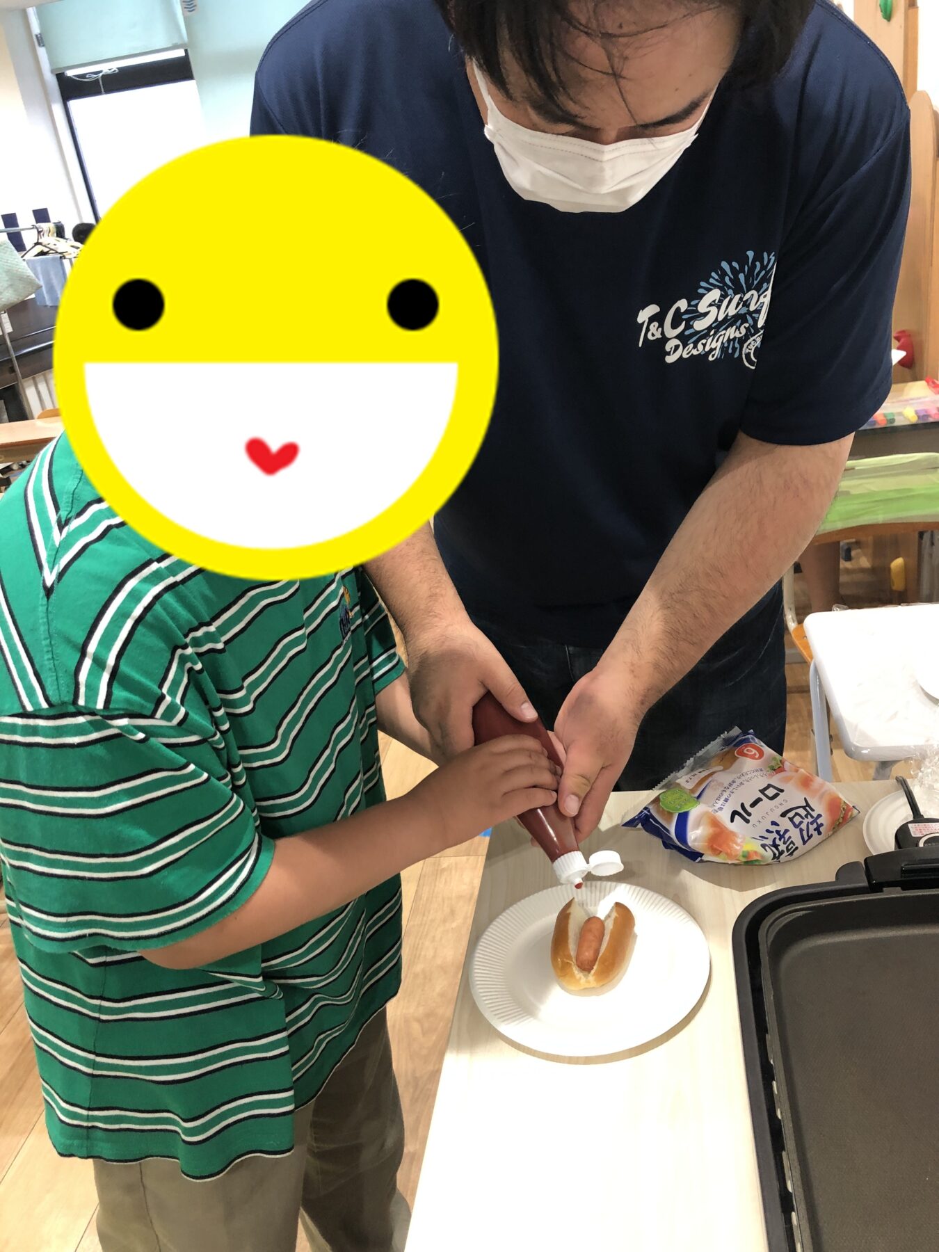 Adult wearing a mask helps a child prepare hot dogs at a kitchen counter, with buns and sausage on the table.