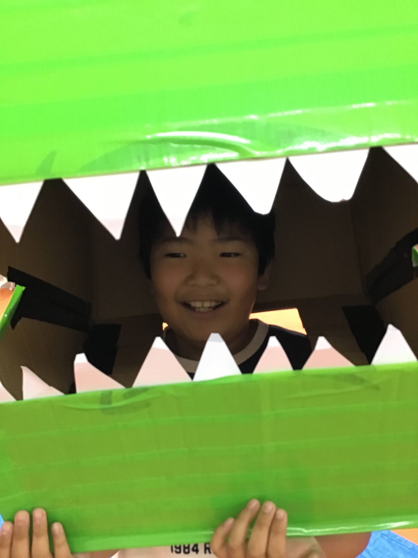 Child smiling inside a green cardboard creature with white triangular teeth surrounding the opening