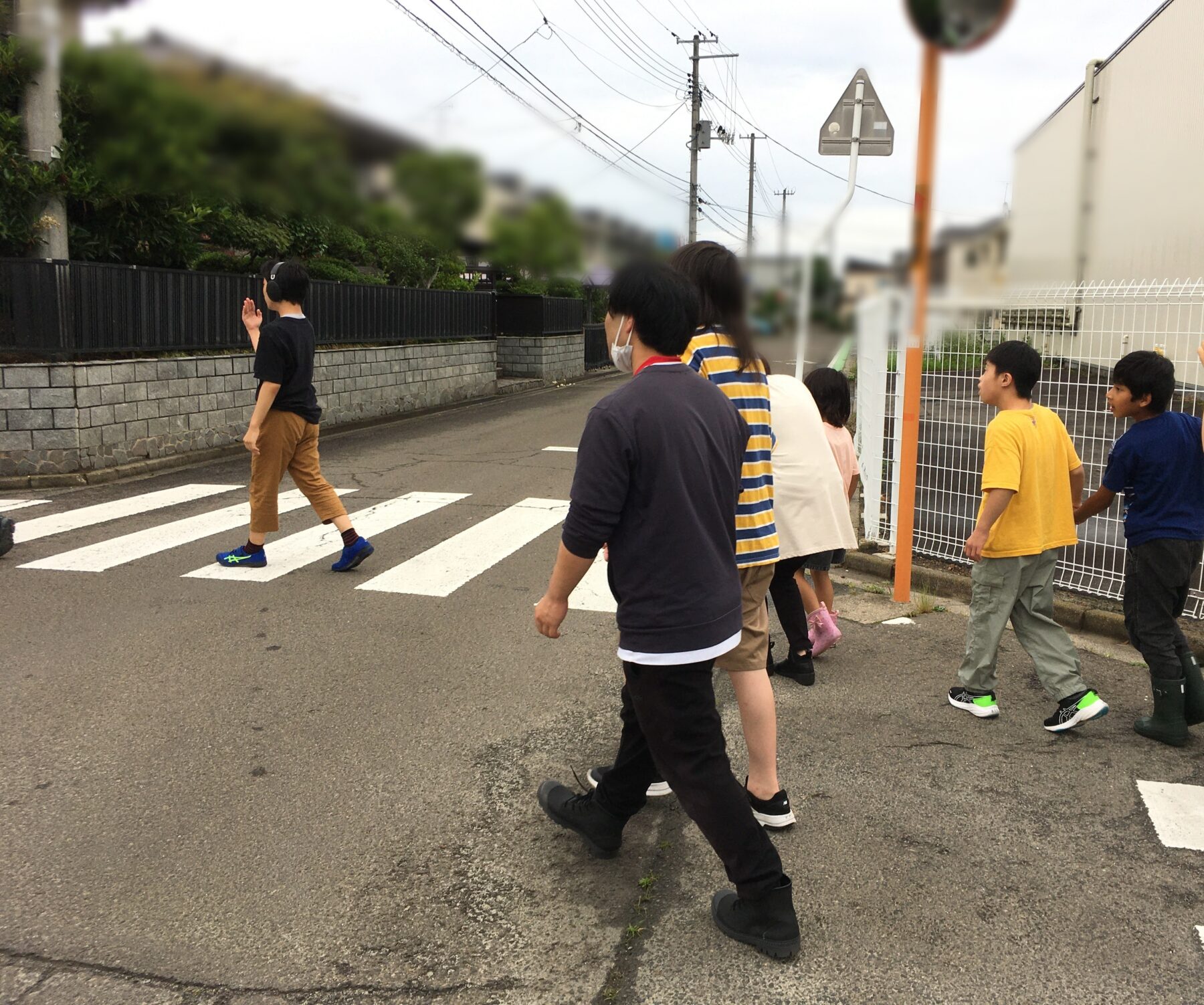 Group of kids walking across a zebra crosswalk on a residential street, with a boy in a black shirt waving as they walk past a fence and power lines.