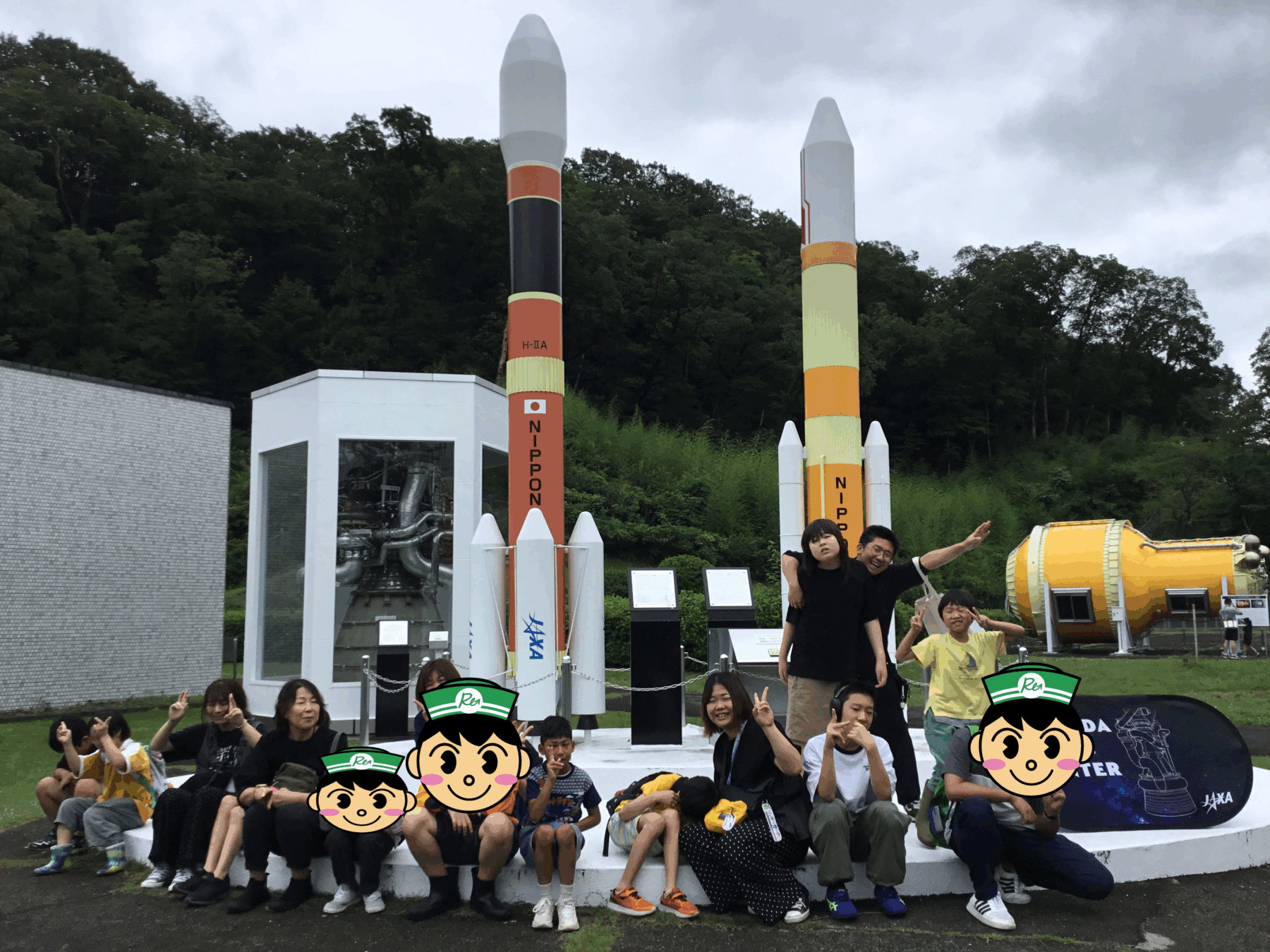 Group of people, mostly kids, sitting on a white platform in front of large model rockets at an outdoor science exhibit; trees and cloudy sky behind them.