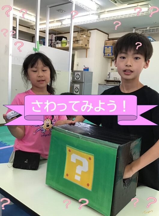 Two kids stand at a table with a green box marked by a yellow question mark; a pink banner in Japanese reads 'Let's touch!' while they look on. (Interactive mystery box activity in a classroom.)