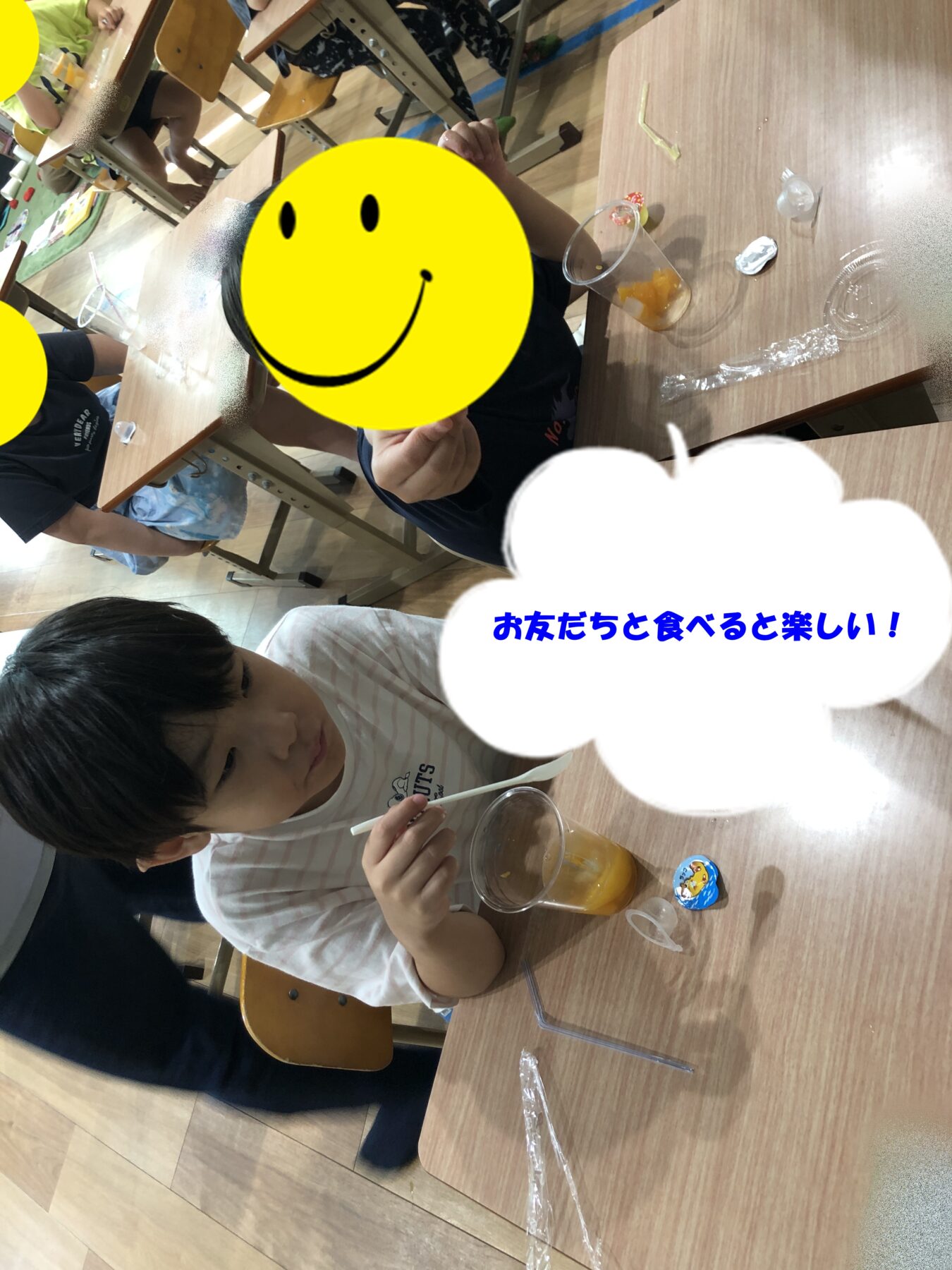 Children sit at desks in a classroom, eating juice and snacks; a large yellow smiley covers one child in the foreground and a speech bubble with Japanese text is visible.
