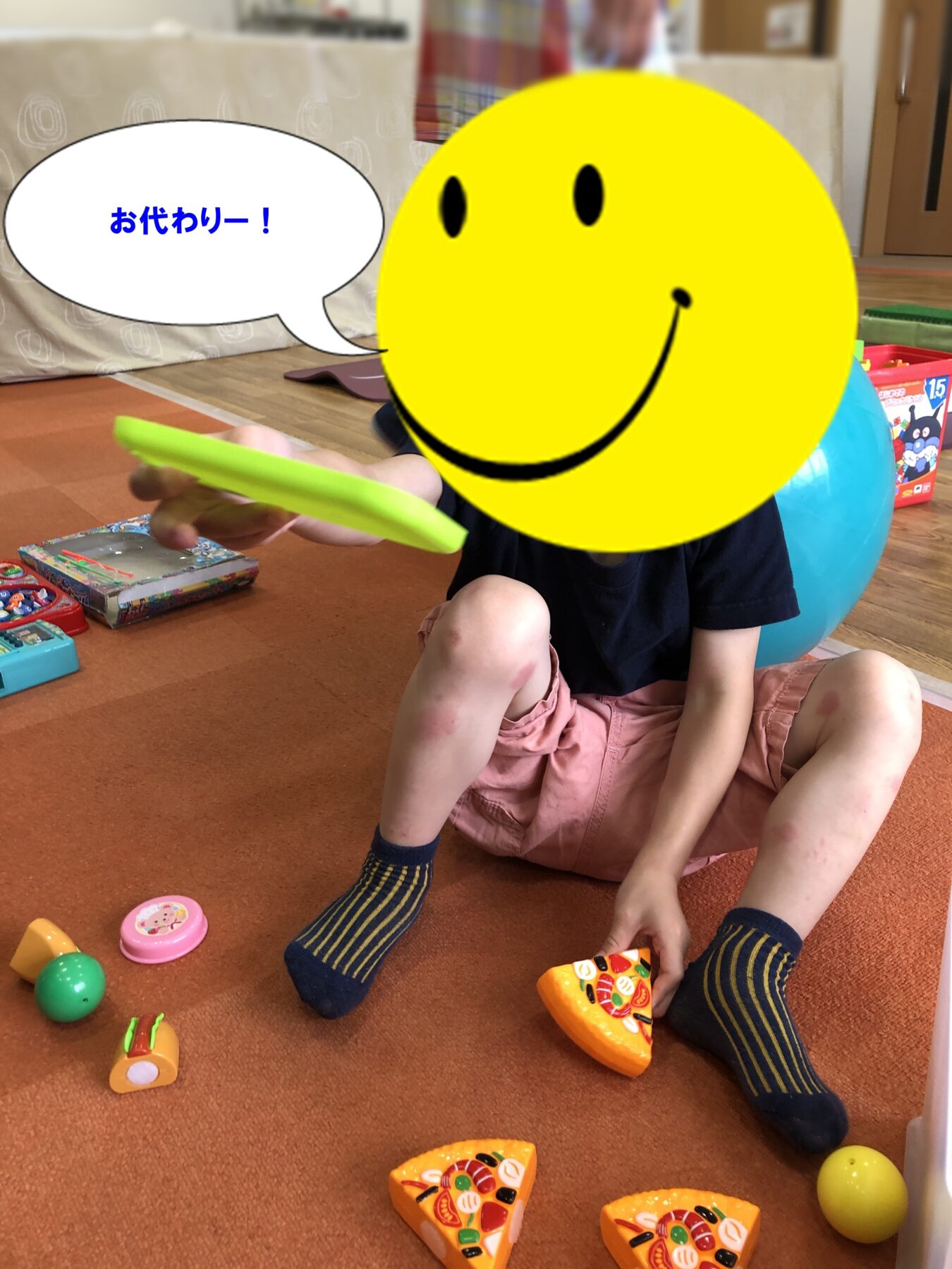 Child sits on an orange carpet playing with toy pizza slices; a large yellow smiley face covers their head and a speech bubble shows お代わりー!