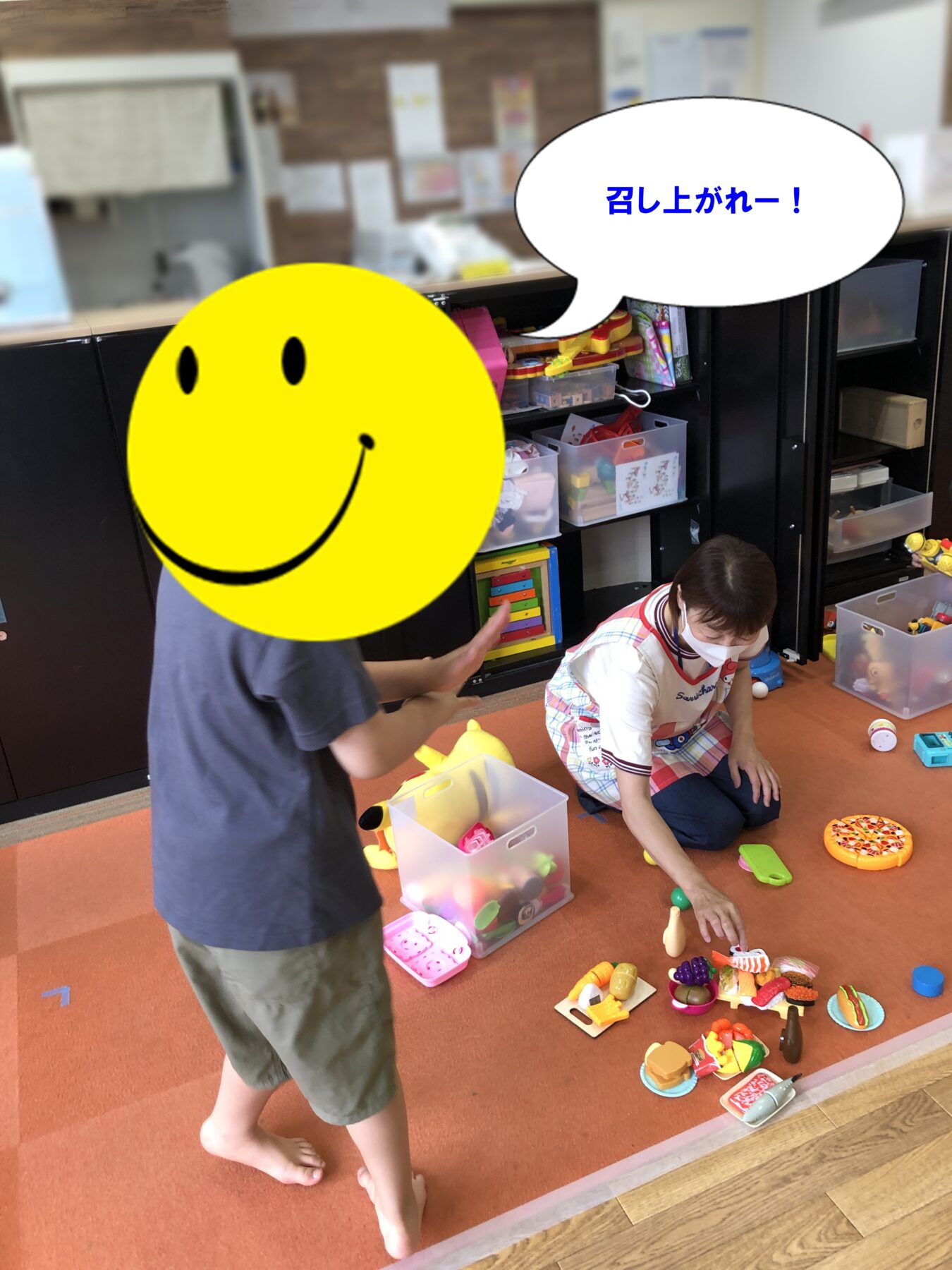 Two children playing with toy food on an orange carpet; one child with a large yellow smiley emoji covering their face, a speech bubble reads “召し上がれー!” (Enjoy your meal).