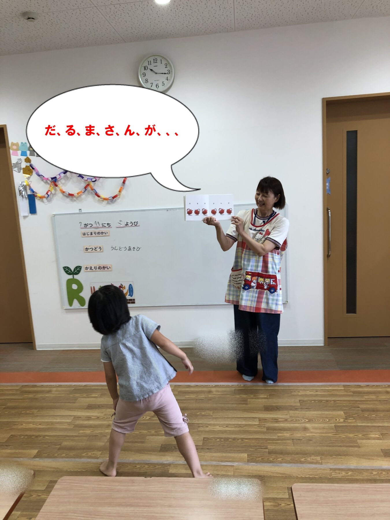 Teacher in a colorful apron shows a card to a seated child in a bright classroom; a large speech bubble with Japanese text overlaps the scene.