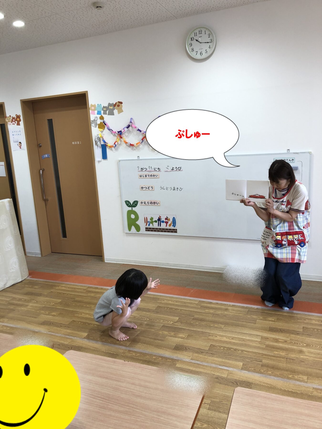 Teacher in a colorful apron shows a picture book to a child on the floor in a bright classroom, with a whiteboard and decorations in the background.