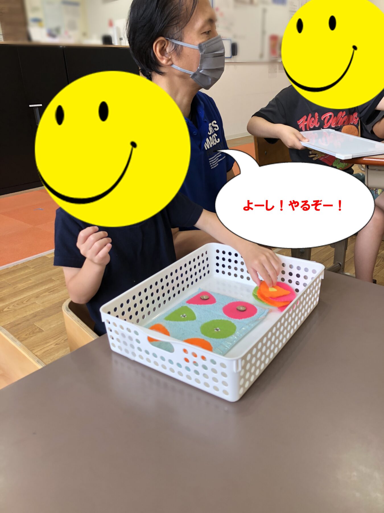 Teacher in a mask helps two children at a table with a colorful water/sensory tray.