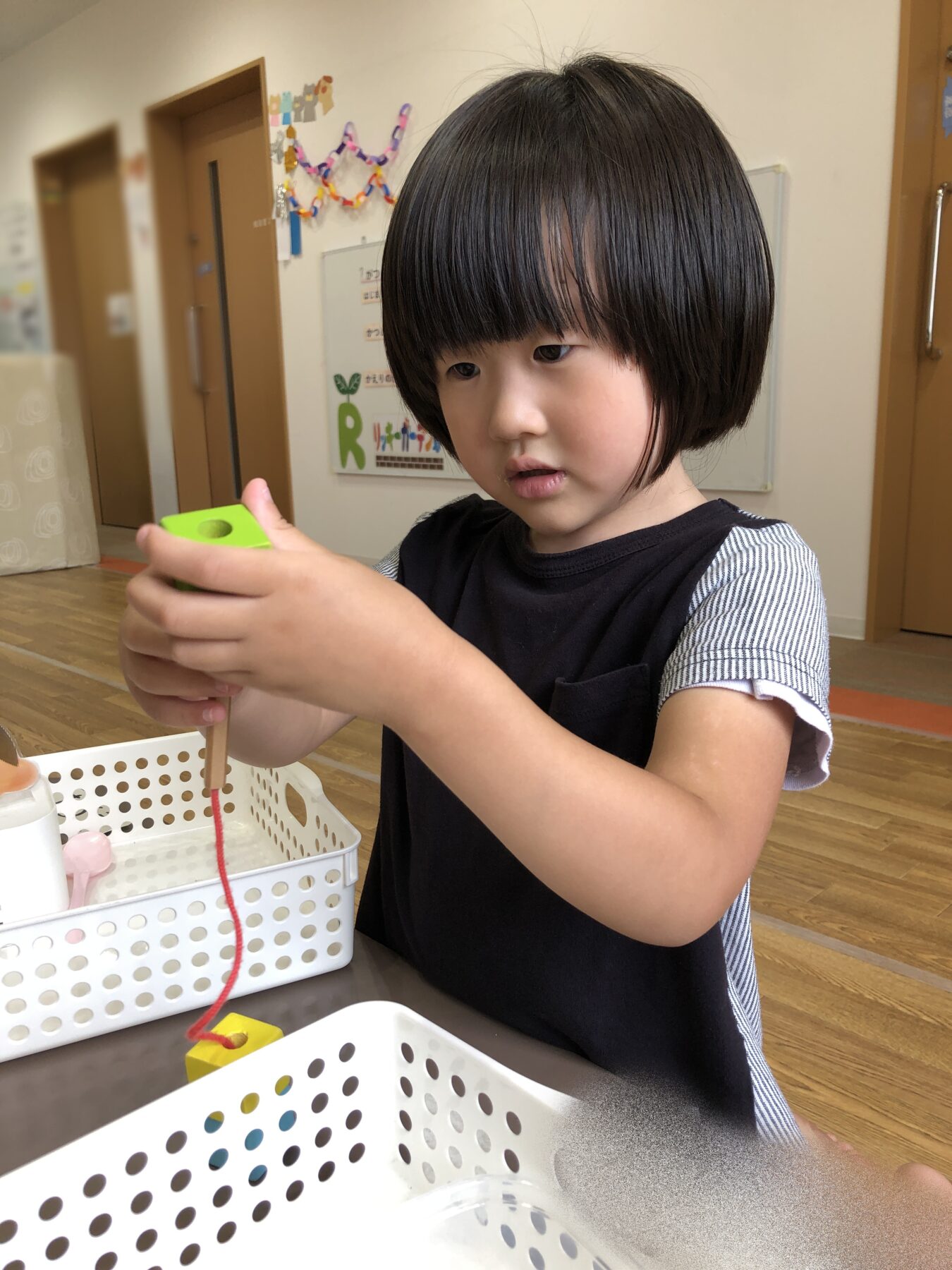 Preschooler concentrates on threading a red string through a green tool at a craft station in a classroom.