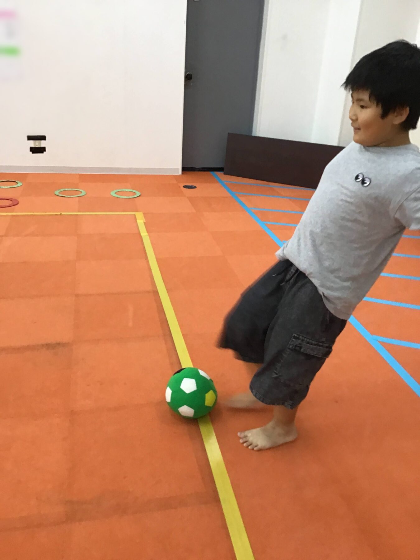 Boy kicking a green and white soccer ball on an orange gym floor with taped lines nearby.