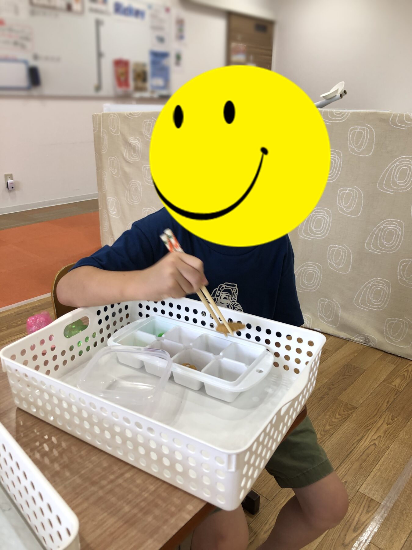 Child with a yellow smiley overlay eating lunch at a desk, using chopsticks over a white lunch basket with divided compartments in a classroom setting.