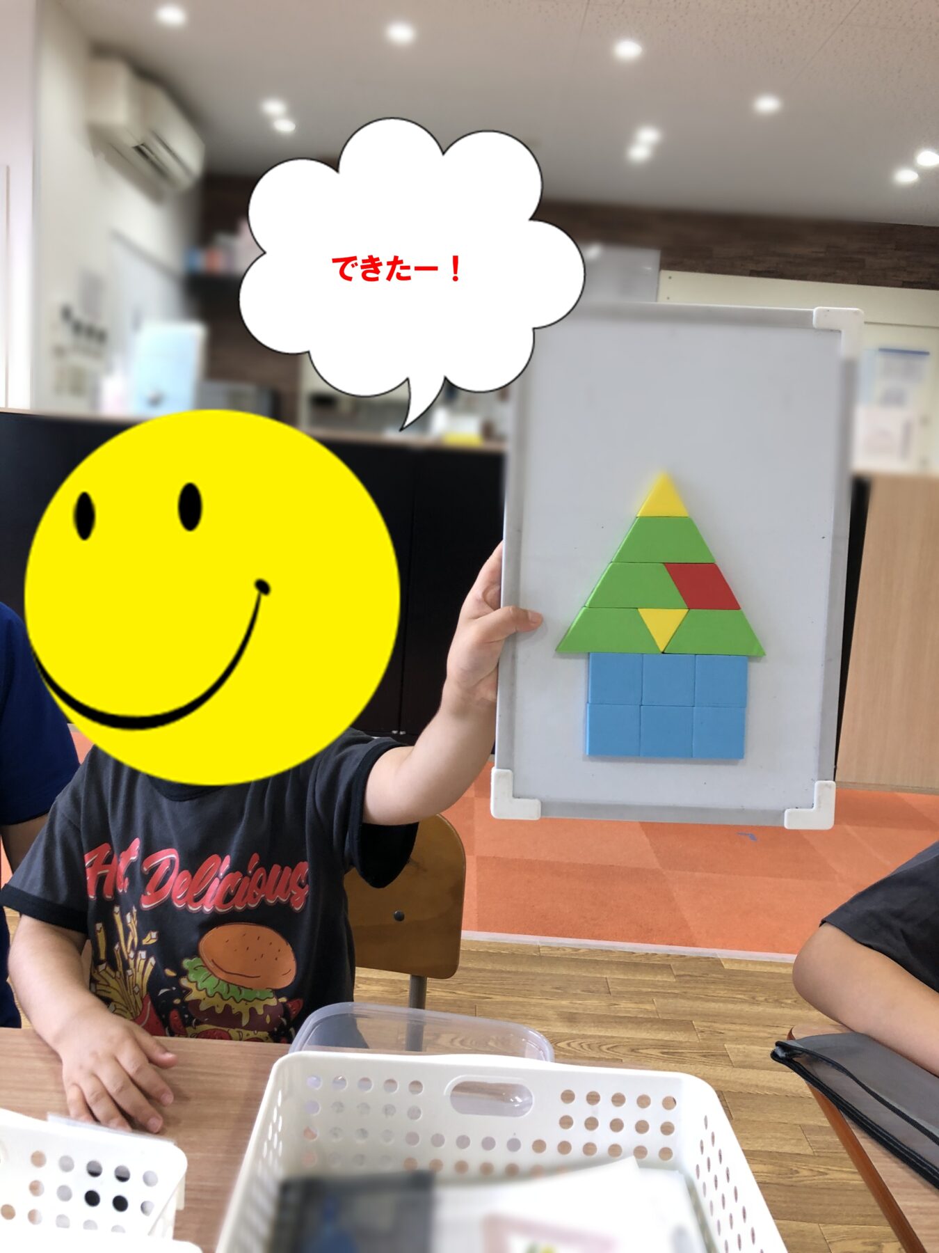 Child presents a clipboard with a completed foam pyramid (green triangles, blue squares) on a blue base; a large yellow smiley mask covers their face, speech bubble reads できたー!