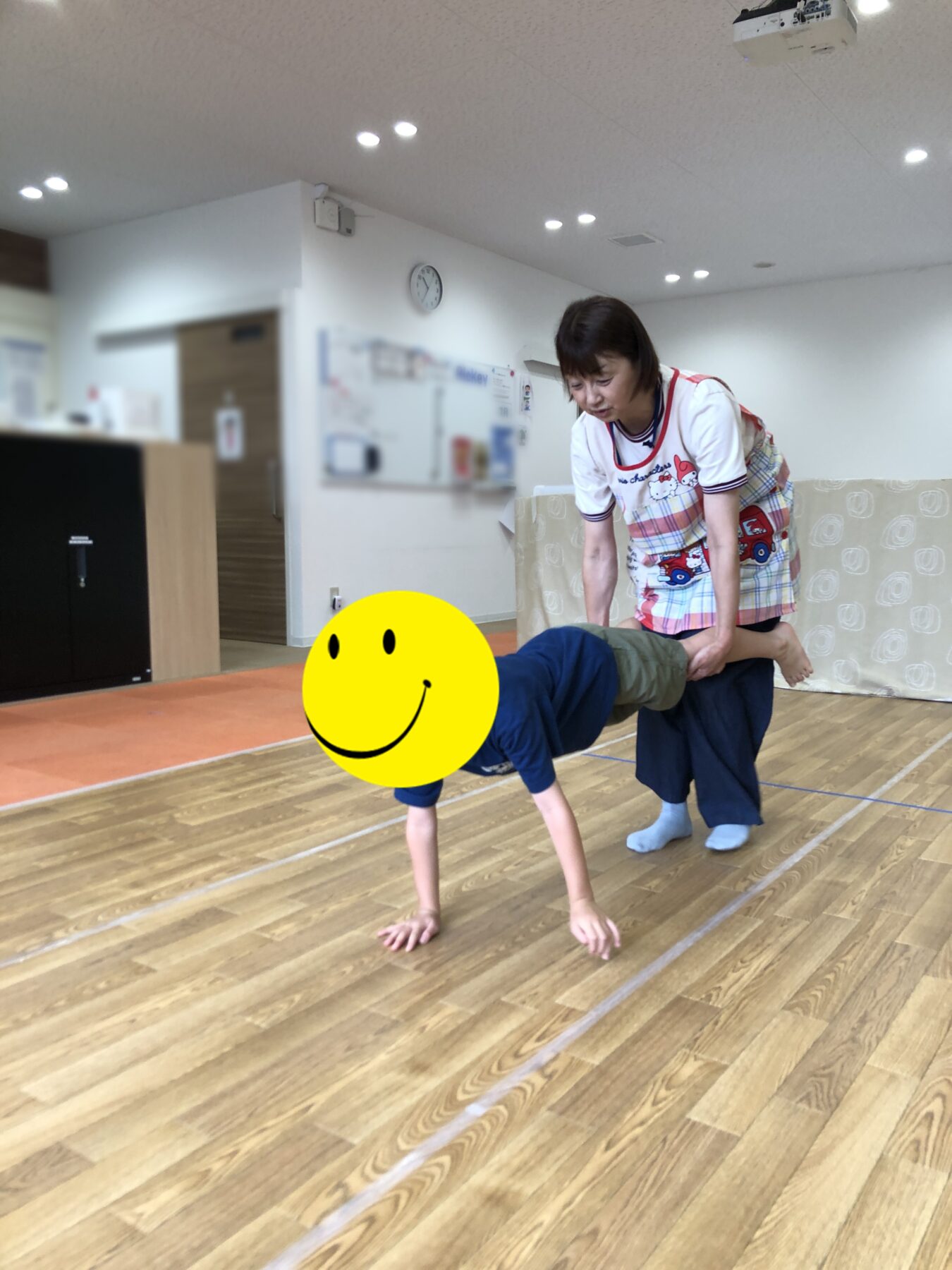 Adult in a colorful apron guides a child doing a wheelbarrow walk in a bright gym. (child's face covered with a yellow smiley sticker)