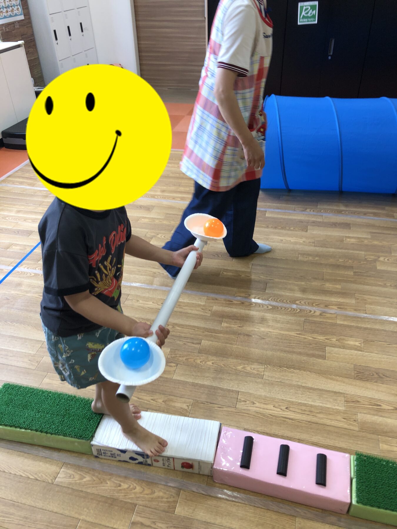 Child playing indoors with a toy bowling ramp, holding a rod with orange and blue balls, walking on a wooden floor near foam blocks and a pink platform; an adult stands nearby in a plaid shirt.