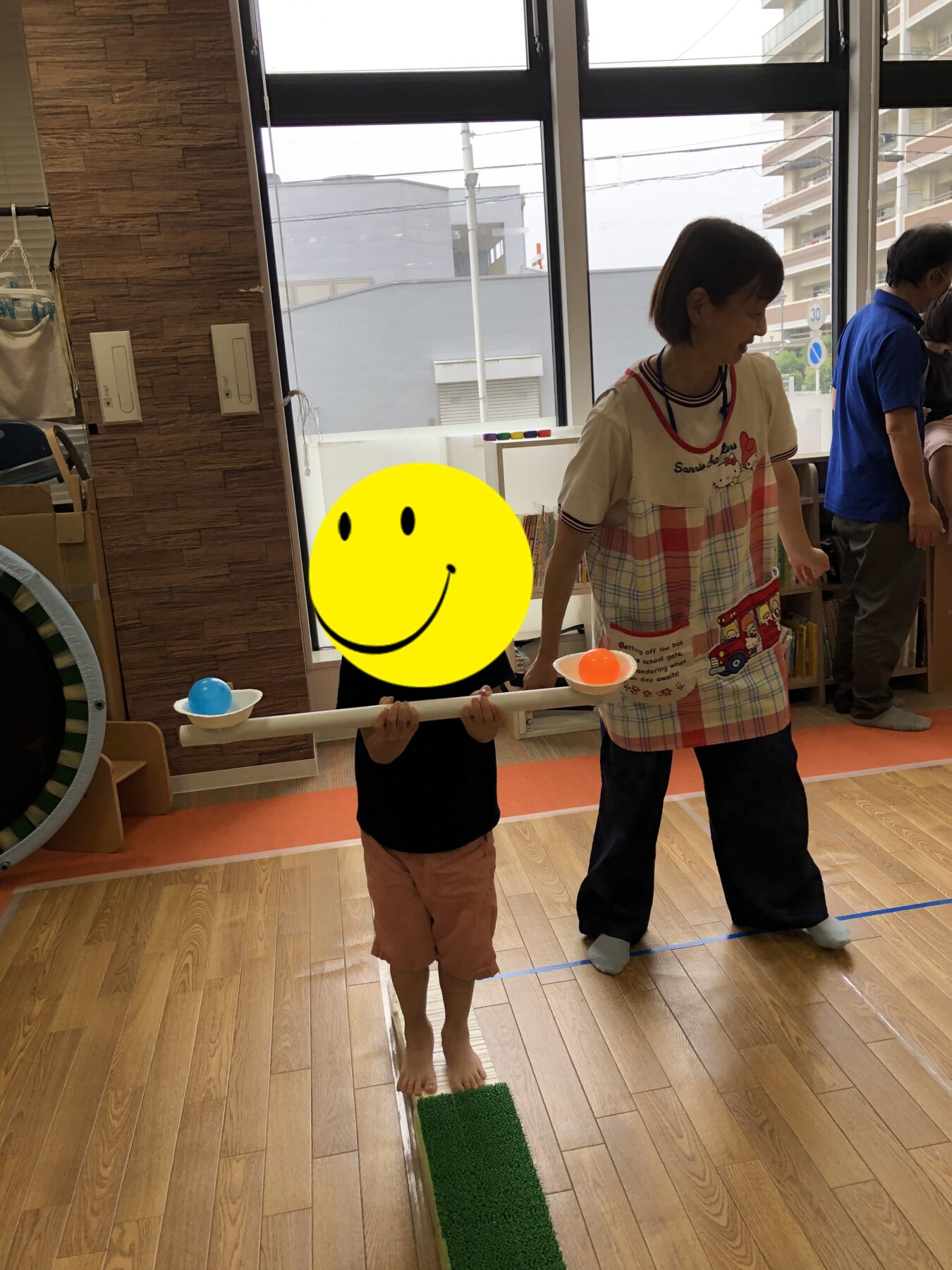 Child in a plaid apron balances on a low beam, holding a long white rod with balls at each end in a classroom play area.
