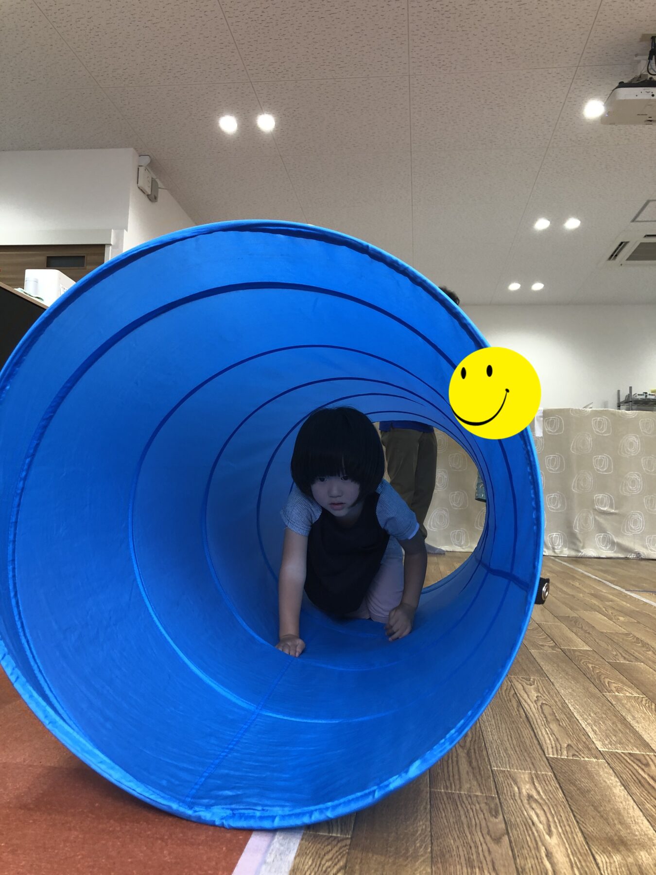 Child crawls through a large blue indoor play tunnel on a wooden floor in a bright room with overhead lights, smiley sticker on the side.