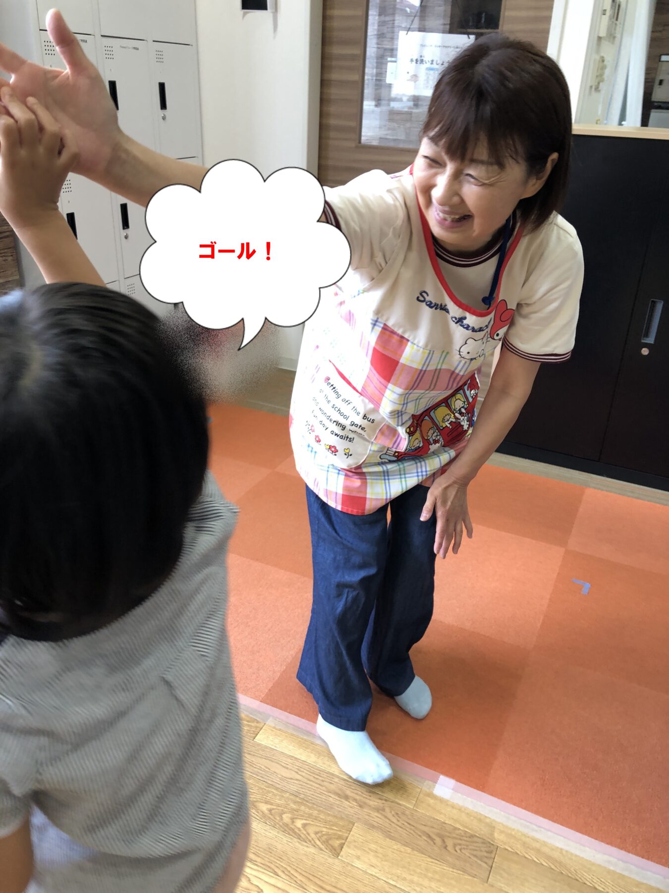 Smiling woman in a white shirt and colorful apron high-fives a child in a room with lockers, a speech bubble reads 'ゴール!' indicating a goal celebration.