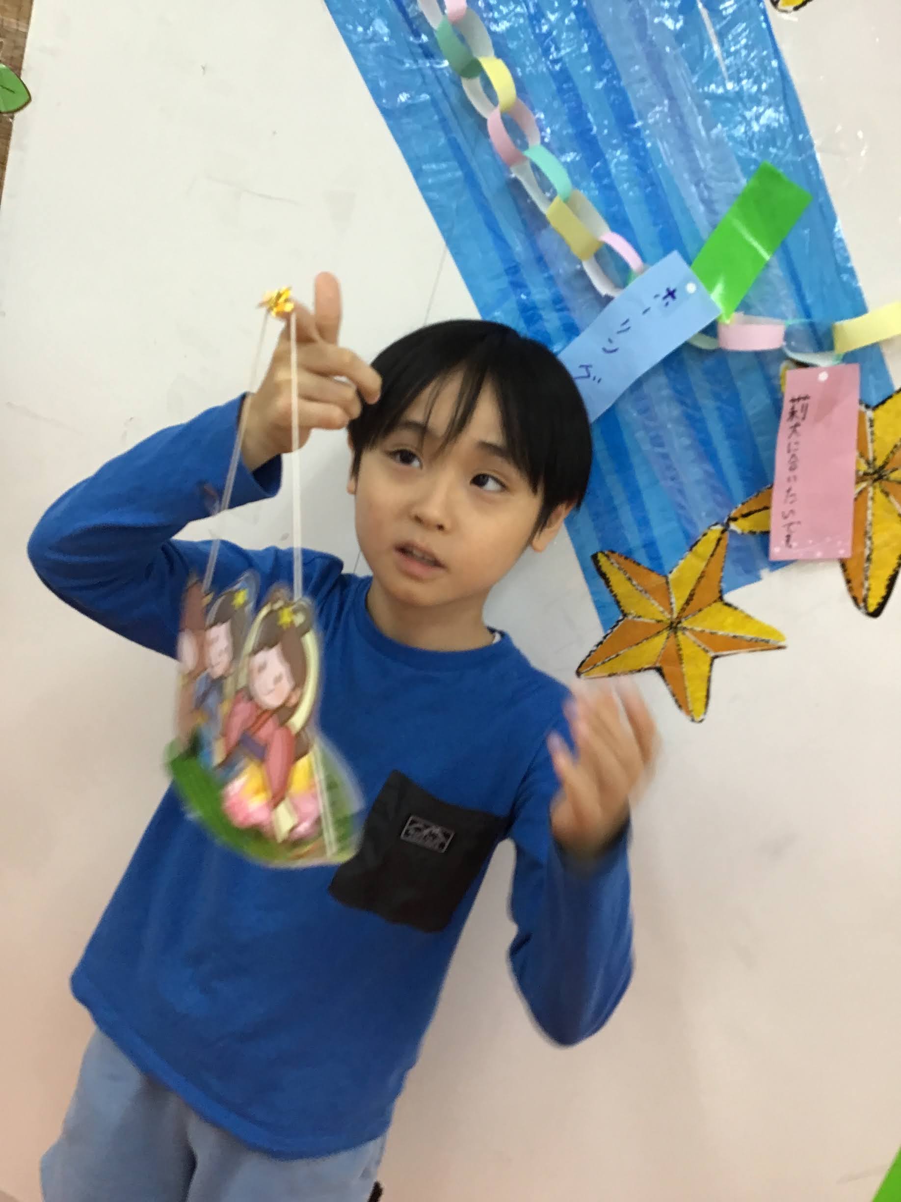 Young boy in a blue shirt holds up a hanging photo craft in front of a wall decorated with paper chains and blue streamers.