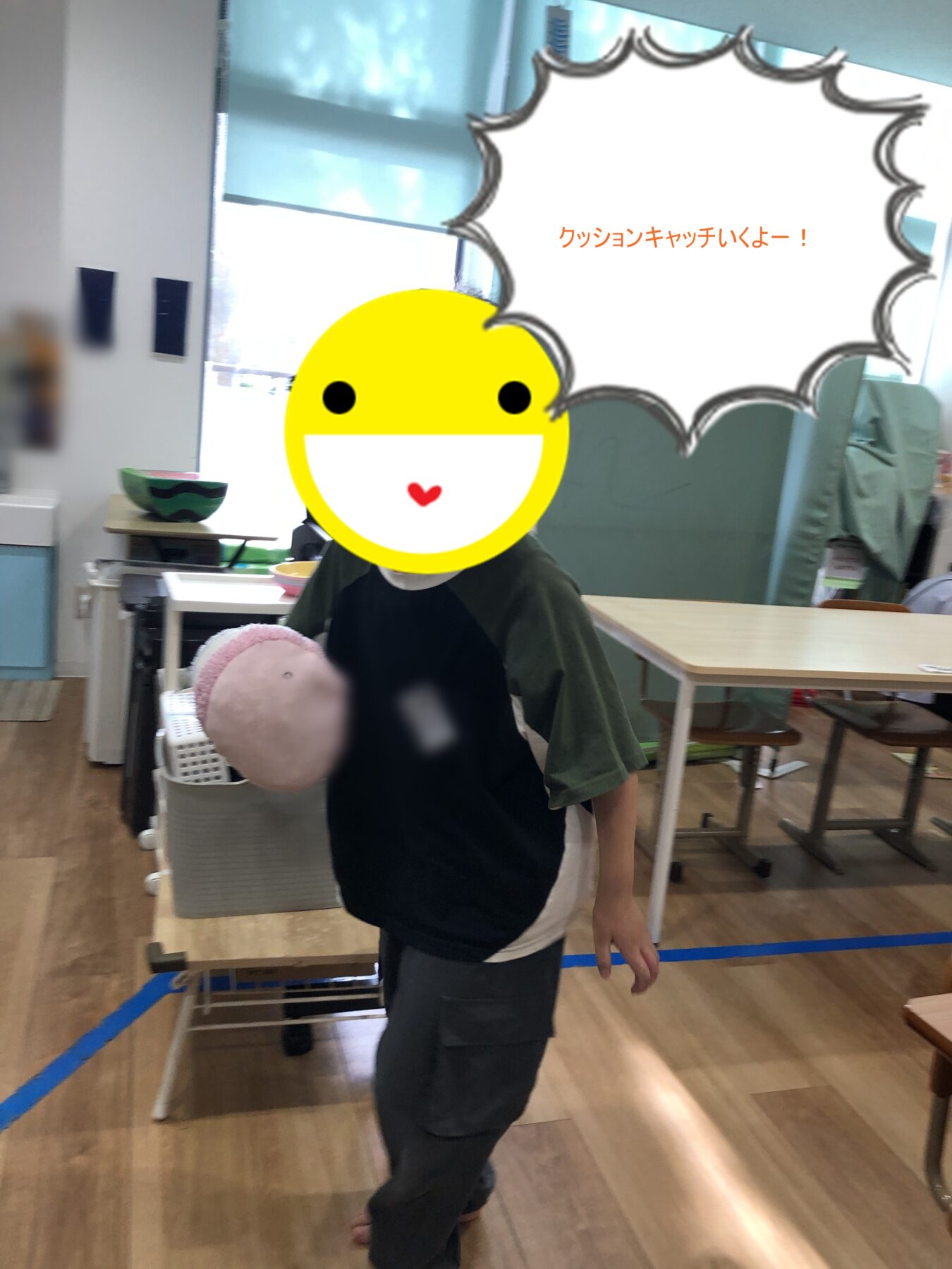 Child in a classroom with tables and chairs, holding a pink cushion toward the camera; a large speech bubble overlay reads 'クッションキャッチいくよー！'
