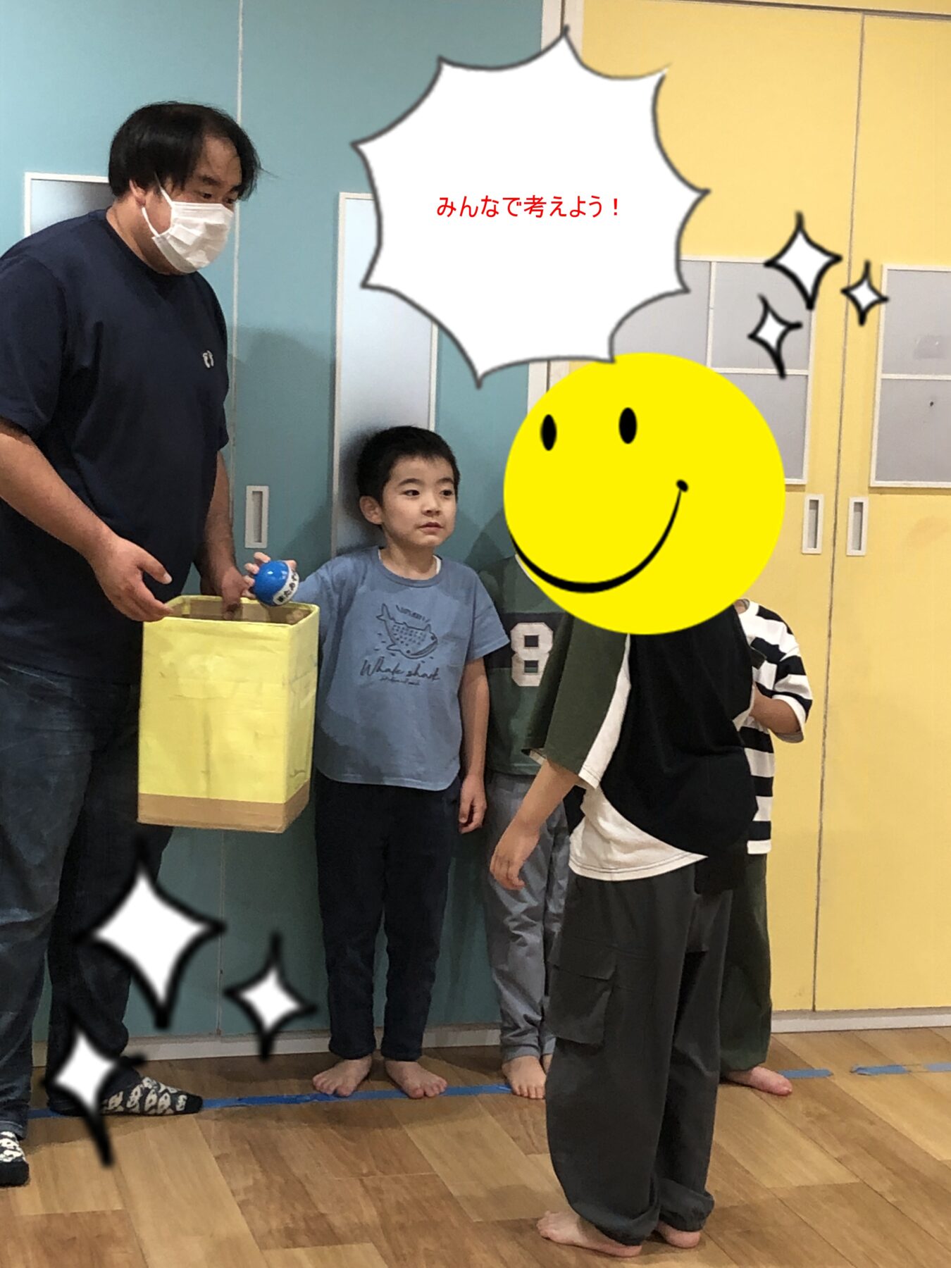 Man wearing a mask hands a yellow box to a standing boy as others watch in a colorful classroom setting.