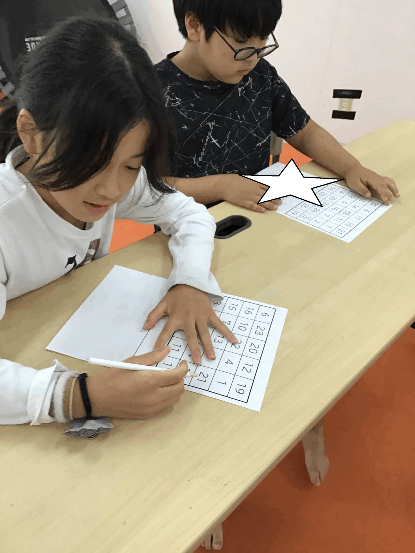 Two children sit at a table solving a number-grid worksheet together in a classroom. One girl writes on a sheet while a boy looks at a nearby grid sheet with a star graphic obscuring part of it.