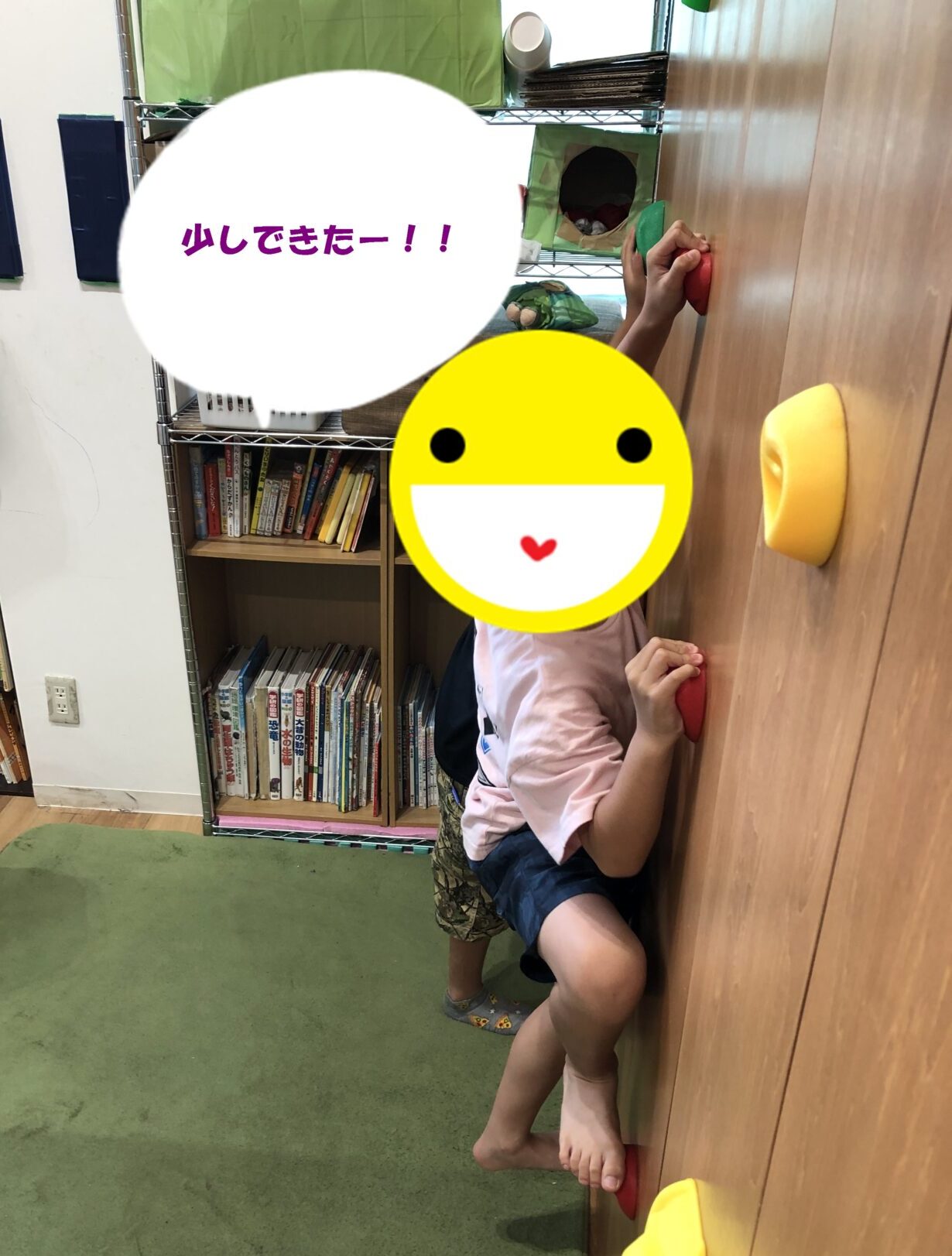 Child climbing a wooden wall with colorful holds in a playroom; a large emoji sticker covers the face.