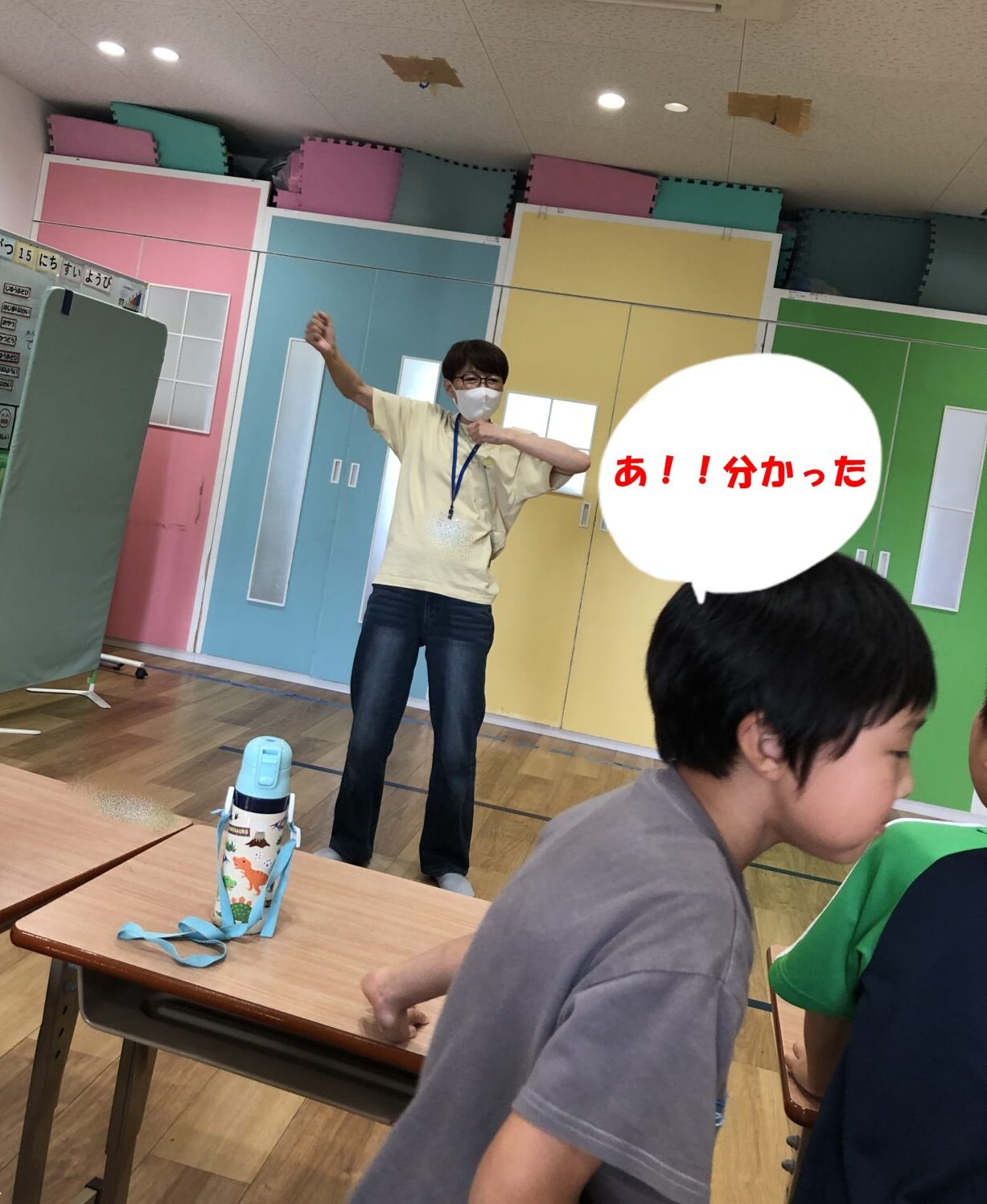 Teacher in a pale yellow shirt and mask gestures with arms raised in a colorful classroom; speech bubble says 'あ!分かった' as students sit at desks nearby.