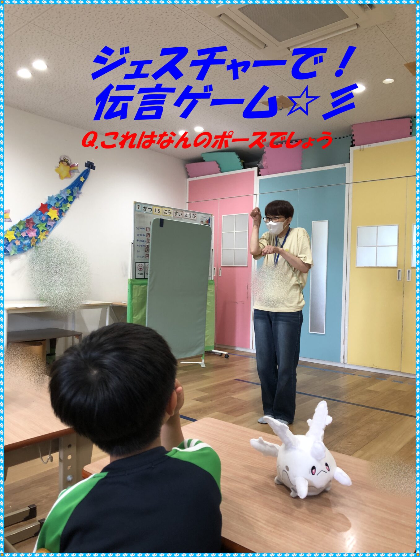 Teacher in a beige shirt and mask leads an activity for attentive children in a colorful classroom; unicorn plush on table nearby.