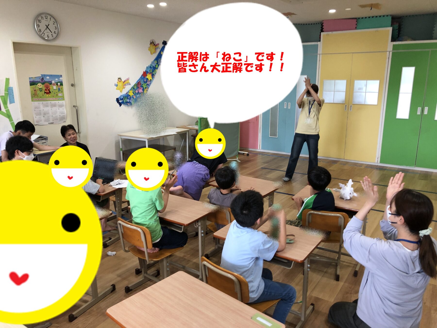 Teacher stands at front of a colorful classroom as students raise hands and look toward the teacher; bright cabinets and desks around. A large speech bubble in Japanese reads: 'Correct answer is