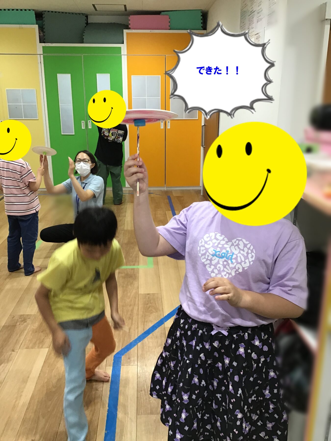 Children indoors playing with spinning plates on sticks as an adult watches; colorful lockers and mats in the background; a speech bubble says 'できた!!'