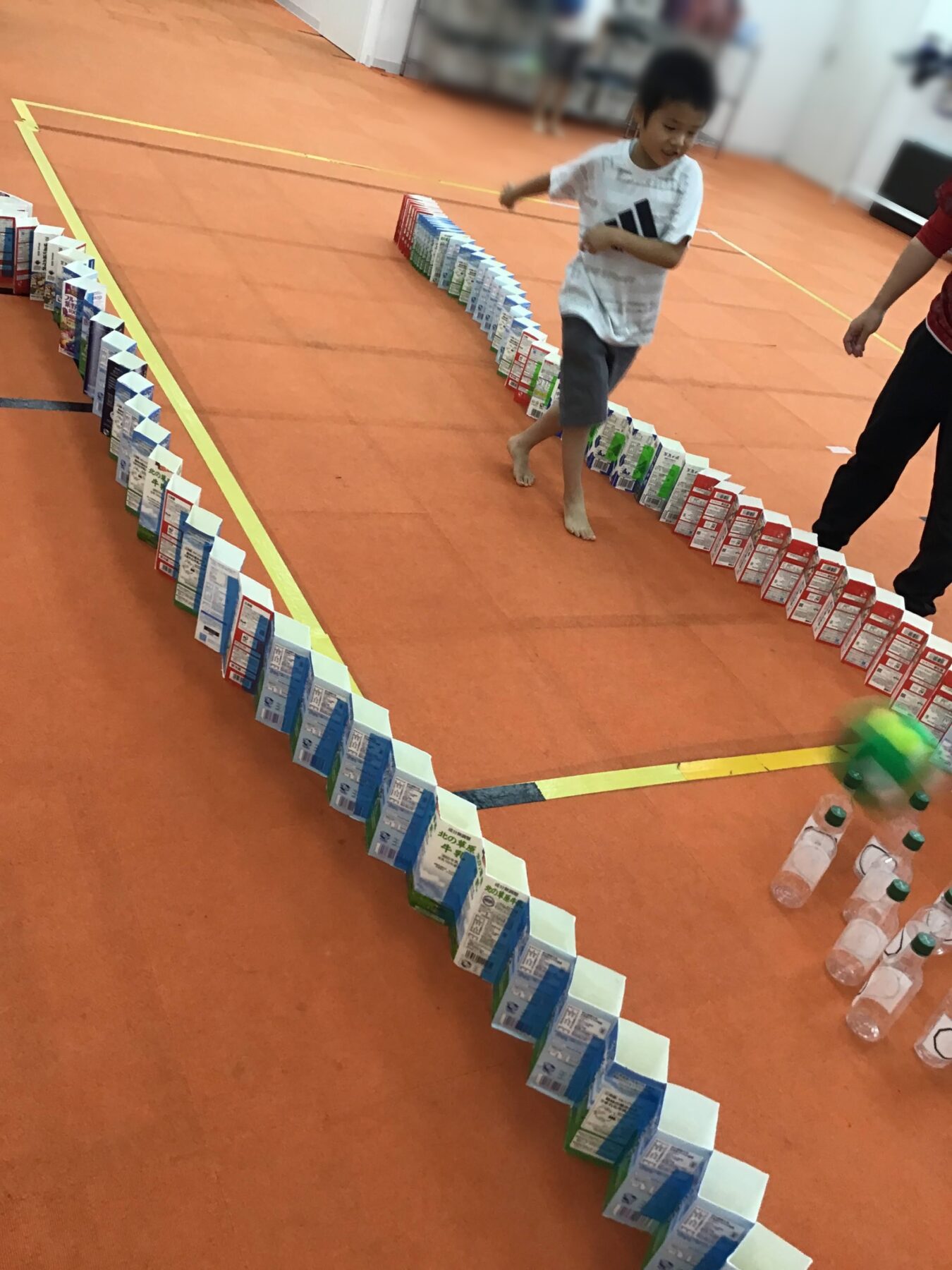 Child walks barefoot along a curved line of stacked juice cartons and boxes on an orange gym floor, game-like setup in a indoor space.