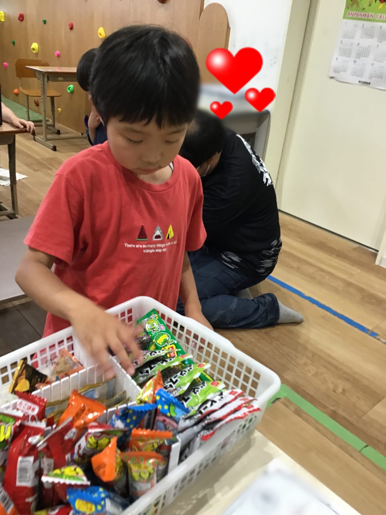 Child in a red shirt selecting candy from a white basket of assorted snacks in a playroom.